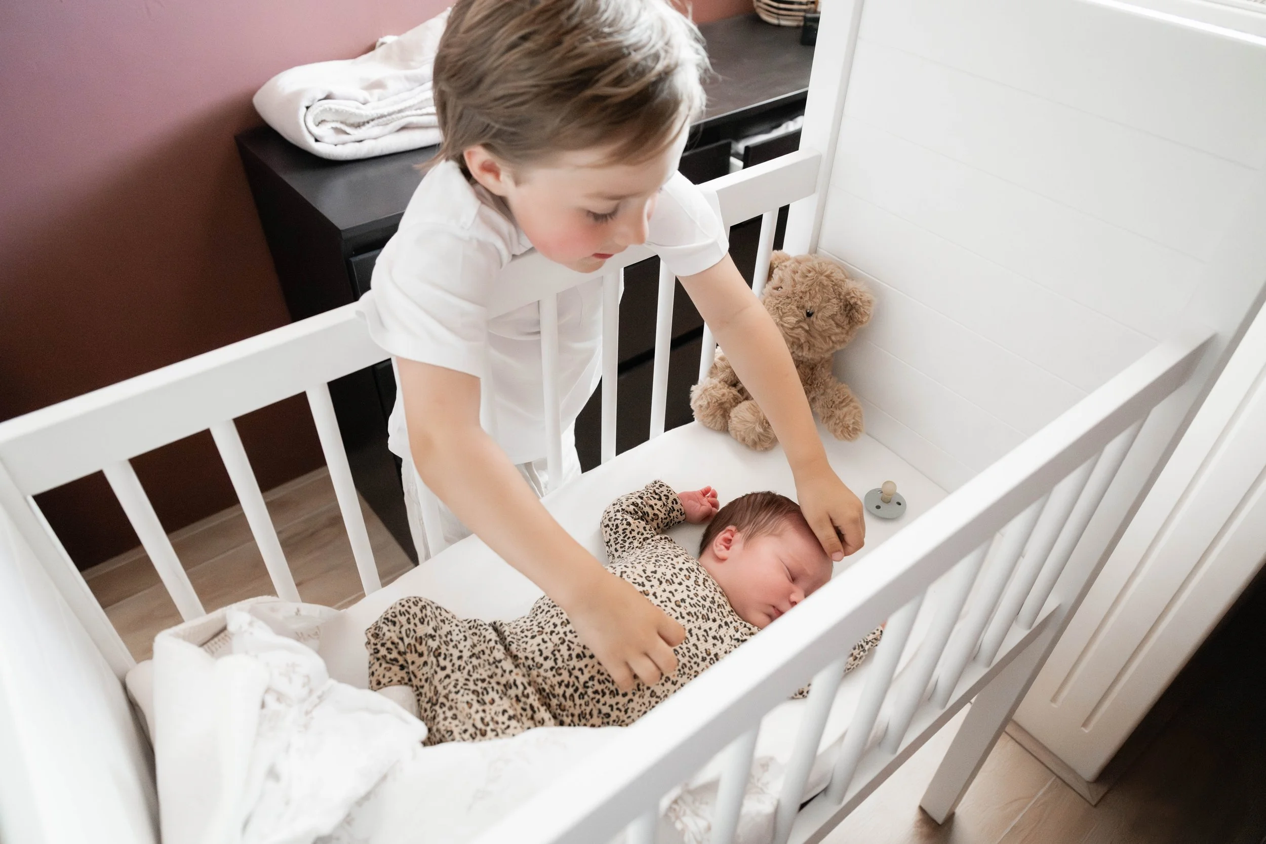 Een oudere kind dat een slapende baby in een witte wieg verzorgt, met een knuffelbeer op de achtergrond.