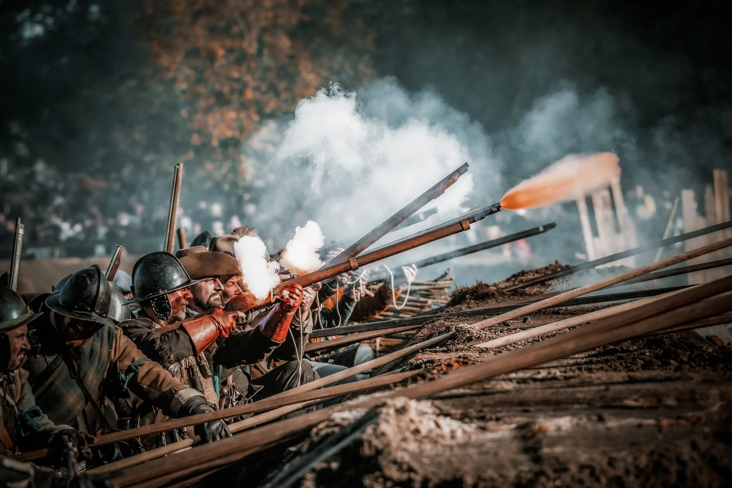 Mensen in historische militaire uniformen met muskets in gevecht, met rook en vlammen die uit de wapens komen, tijdens een oorlogssimulatie of herdenkingsgebeurtenis.