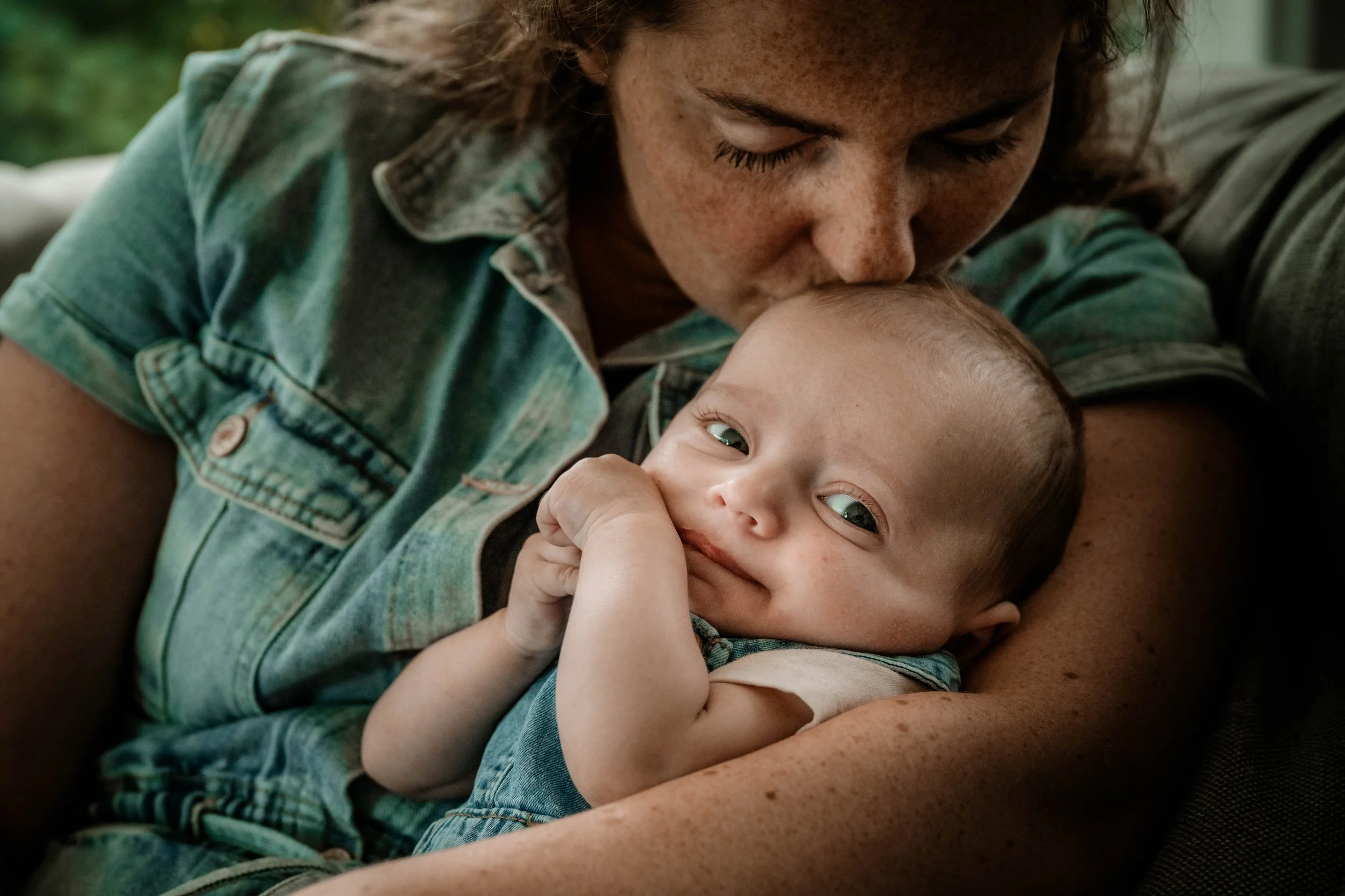 Een persoon met gekruld haar zoent een baby op het voorhoofd, terwijl de baby in iemands armen ligt en naar de camera kijkt.