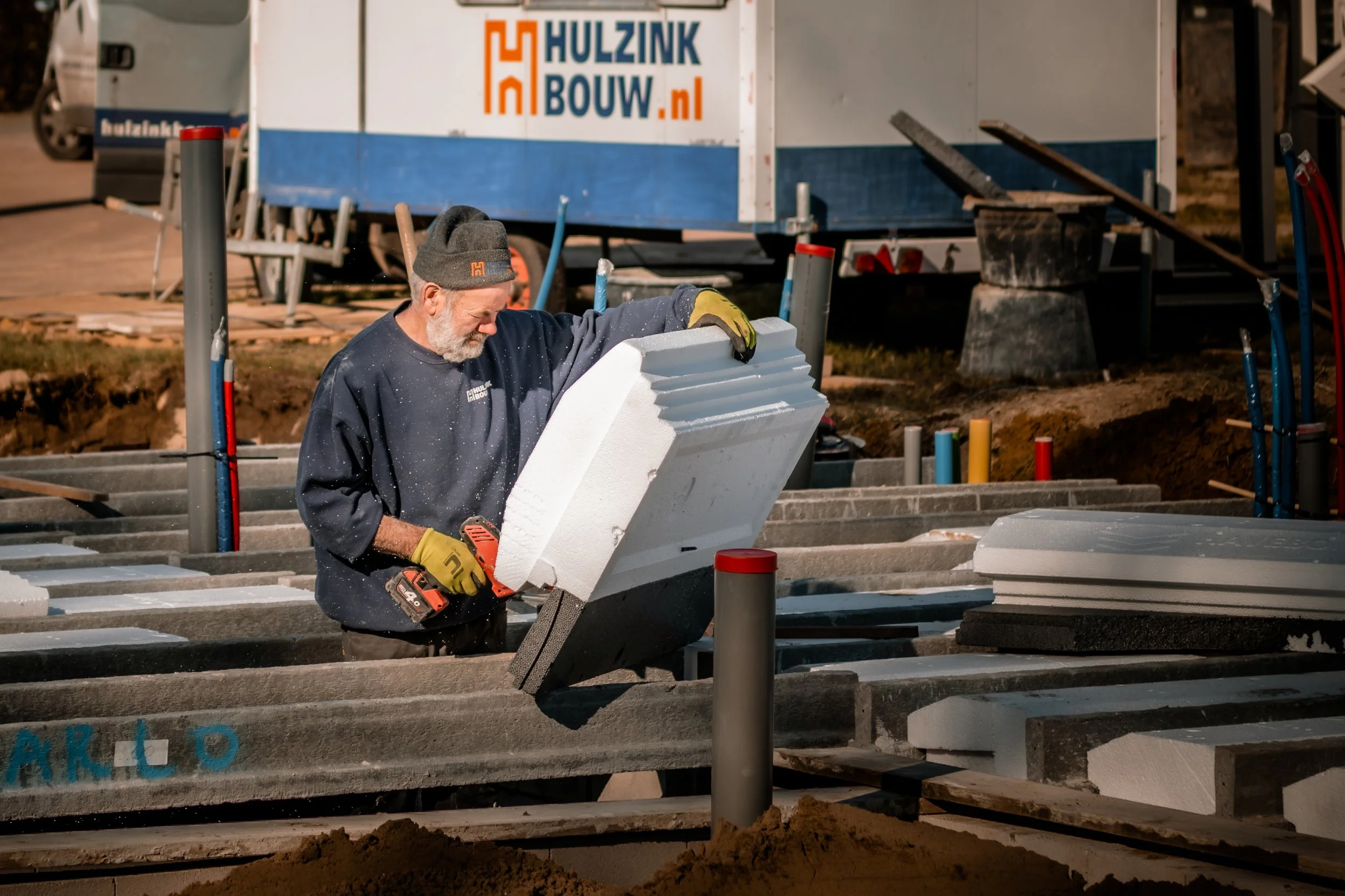 Man werkt met schuimisolatie op een bouwplaats, omringd door bouwmaterialen en gereedschap.