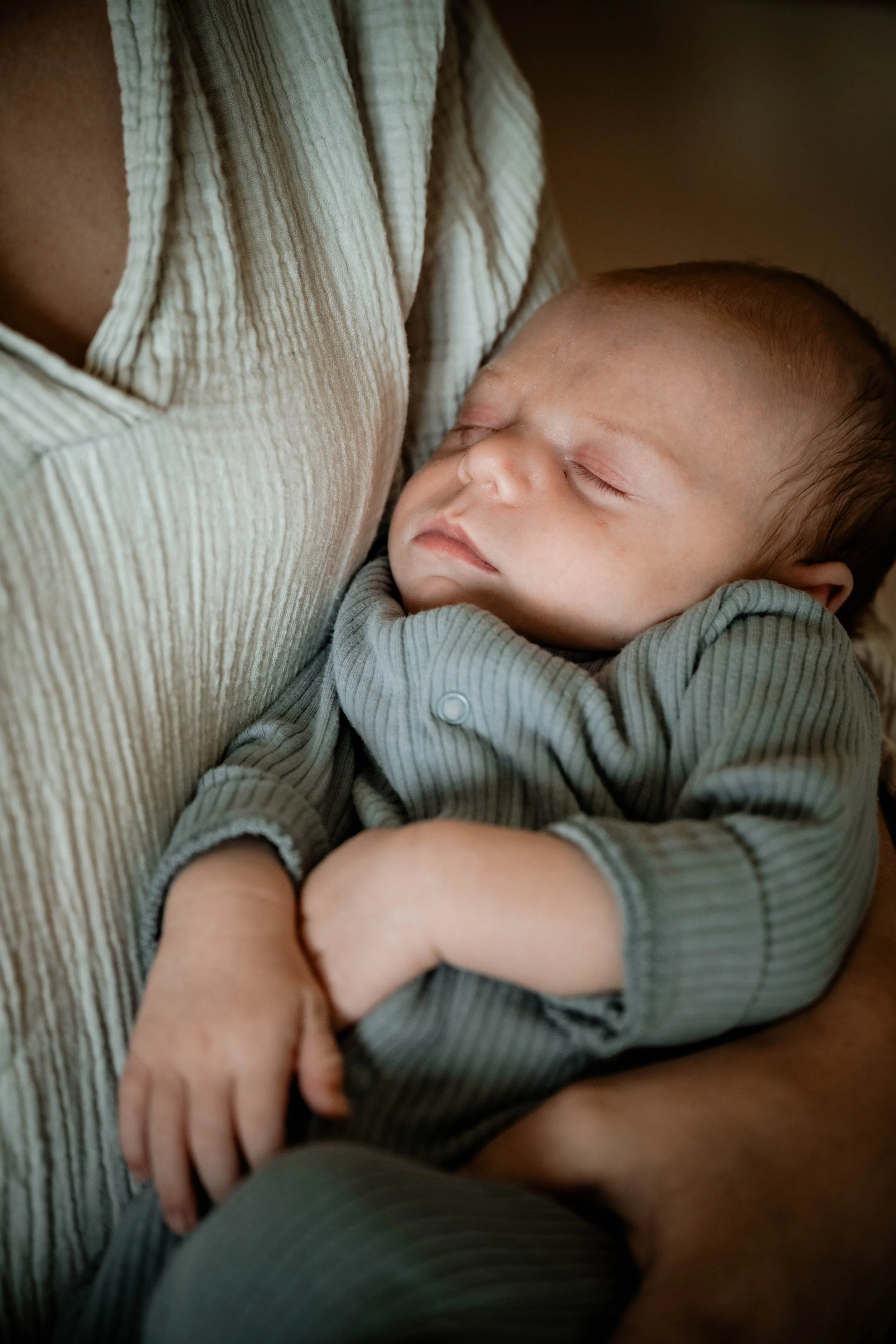 Een baby die vredig slaapt terwijl hij in de armen van een volwassene ligt, gekleed in een grijs geribbelde pyjama, met gesloten ogen en een rustige gezichtsuitdrukking.