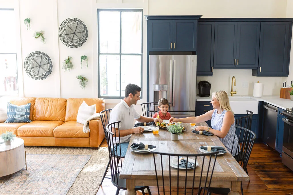 Family sitting at a dining table in a modern kitchen with blue cabinets, stainless steel refrigerator, and large window, enjoying a meal together.