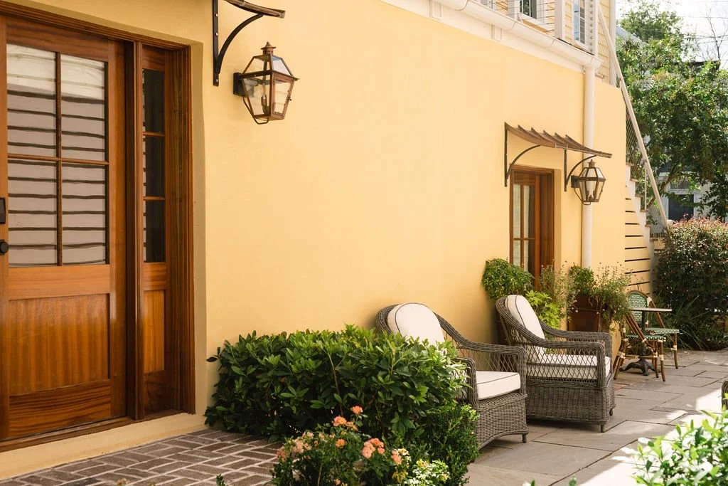Modern house with wooden exterior walls facing a mountain landscape with tall, rugged mountains in the background. There are two people visible inside the house, seen through the large glass window.