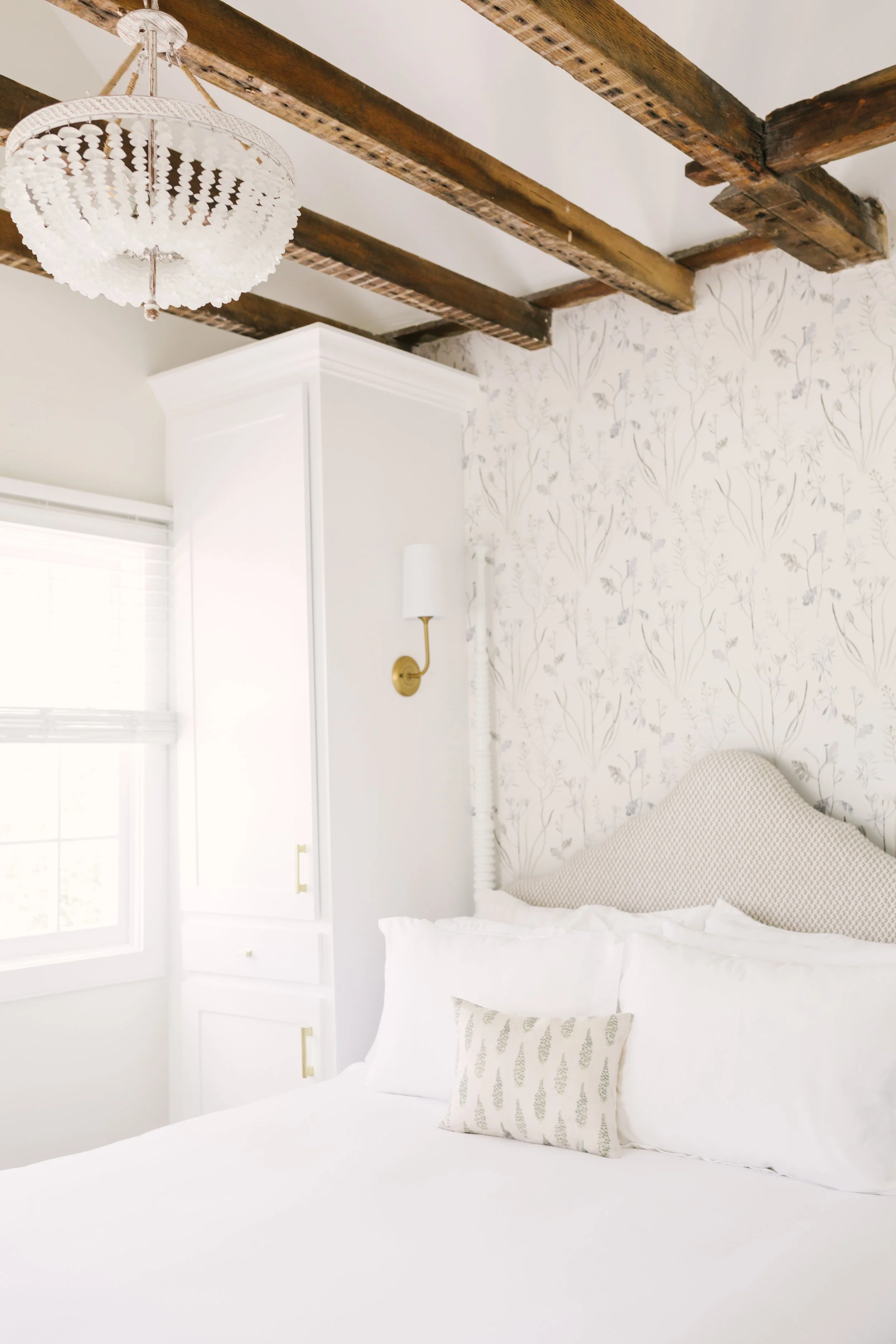 A bedroom with a white bed, decorative pillows, a white wallpaper with a floral pattern, a white wardrobe, a window with blinds, a wall-mounted light fixture, a brass wall sconce, a wooden ceiling with exposed beams, and a white chandelier.