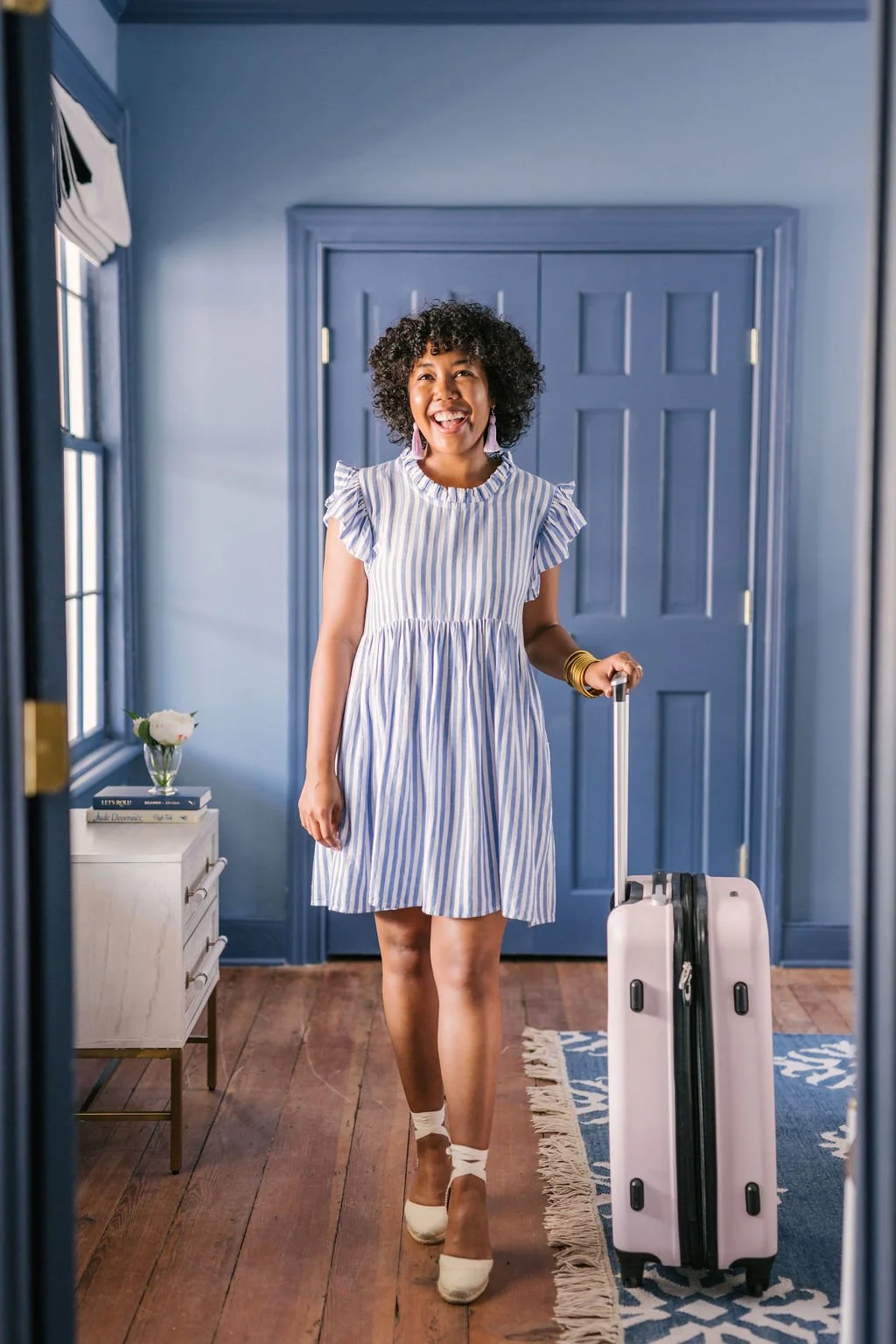 A woman with curly hair smiling and walking with a suitcase in a blue-themed room.