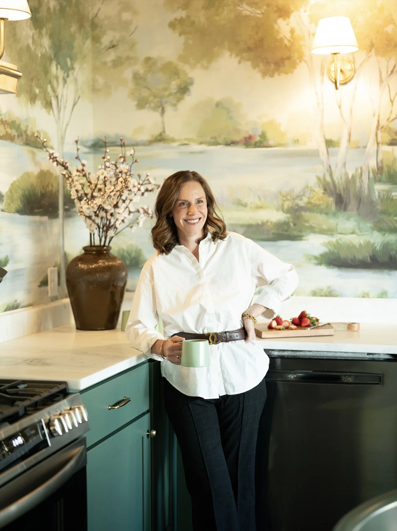 Person placing a book on a round coffee table decorated with a succulent plant, a large wooden vase with branches, and a string of beads, with a patterned rug and window in the background.