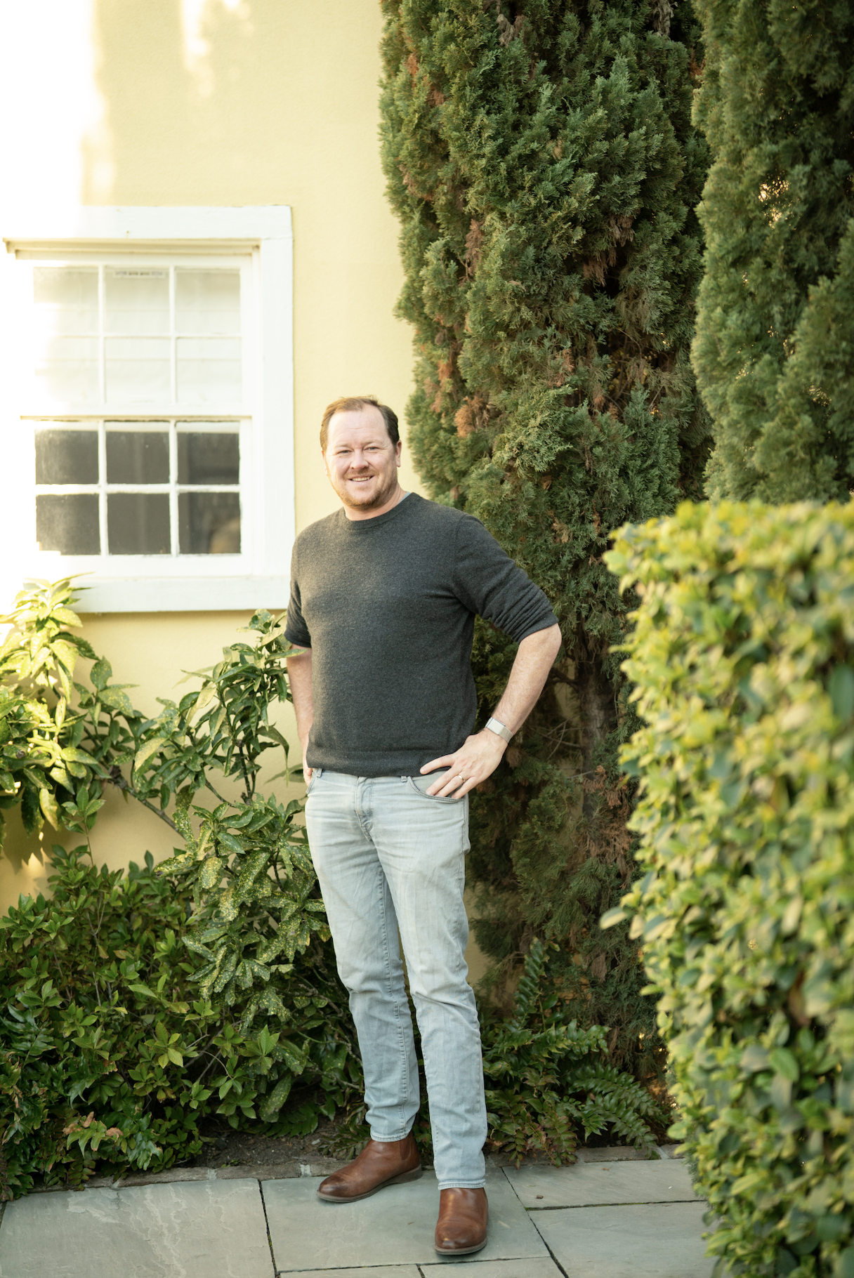A man with short brown hair, glasses, and a big smile, standing in a bright room with white walls. He is wearing a gray knit sweater over a collared shirt.