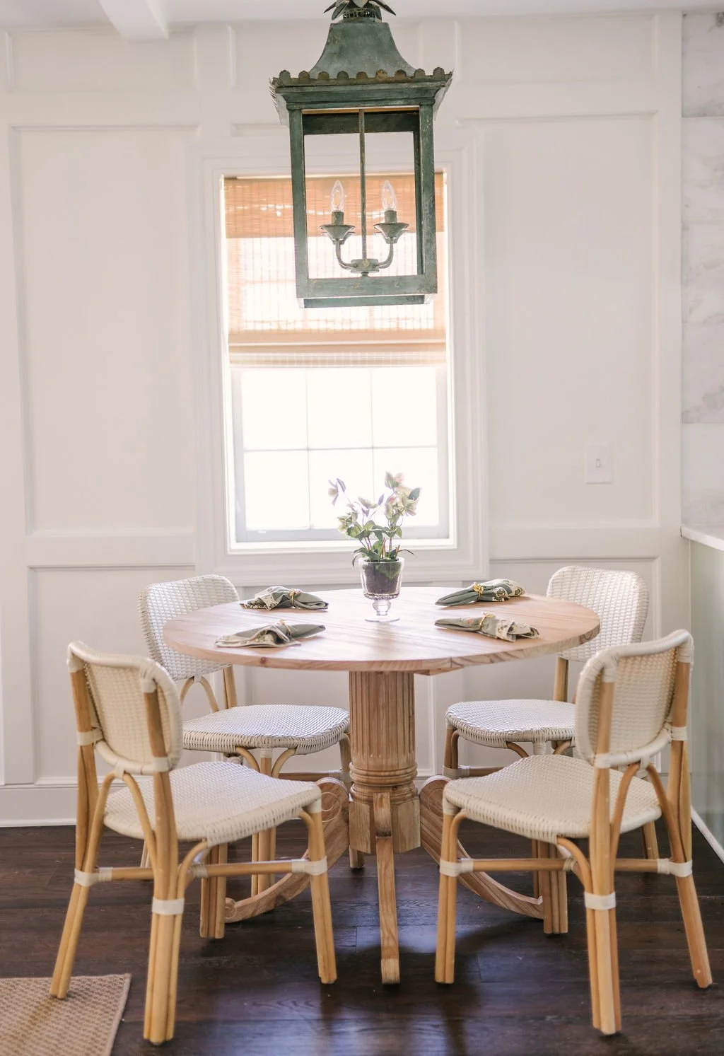 A round wooden dining table with a potted plant in the center, surrounded by five white wicker chairs, and a large window with a bamboo shade in the background, with a vintage metal chandelier hanging above.