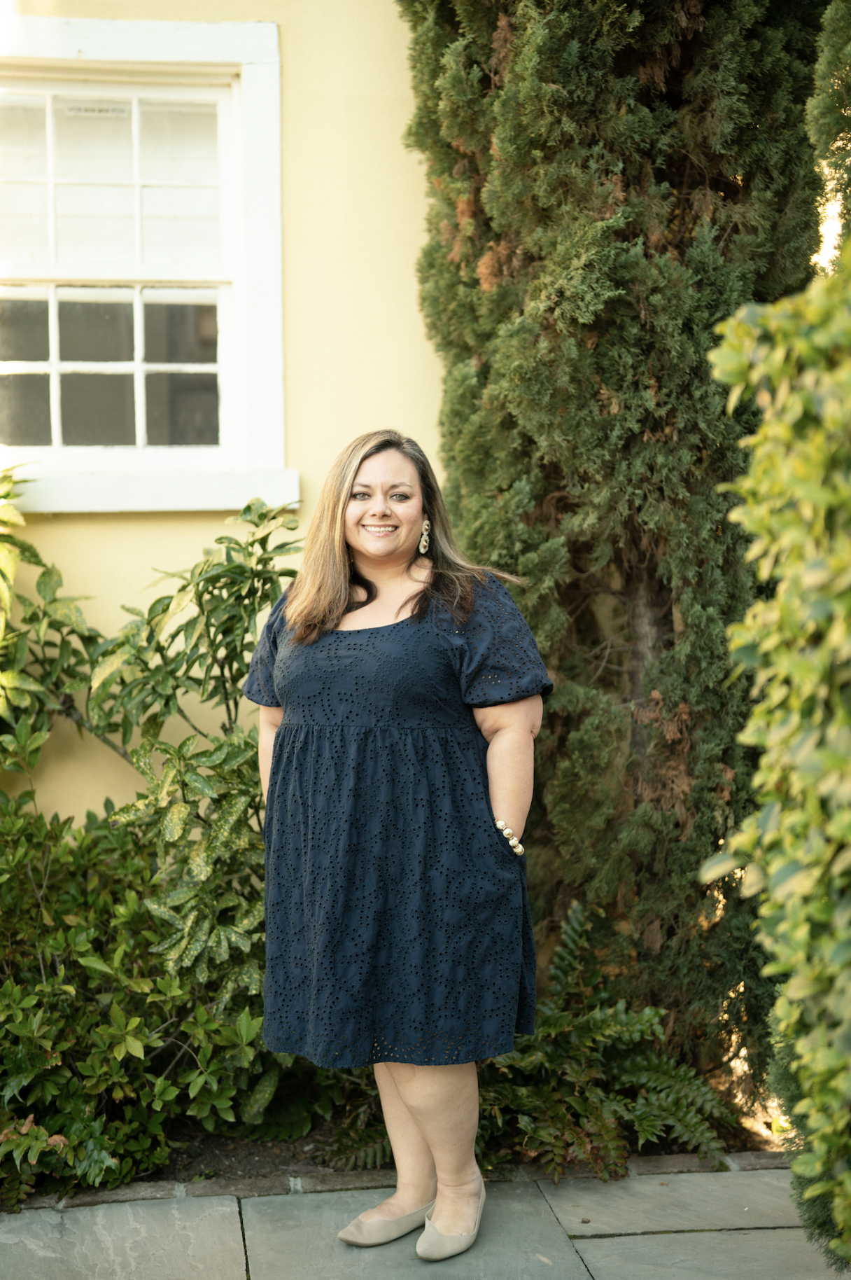 A neatly made bed with white bedding, three brown pillows, a beige throw blanket, a window with white blinds, and two woven pendant lights hanging from the ceiling in a bright bedroom.