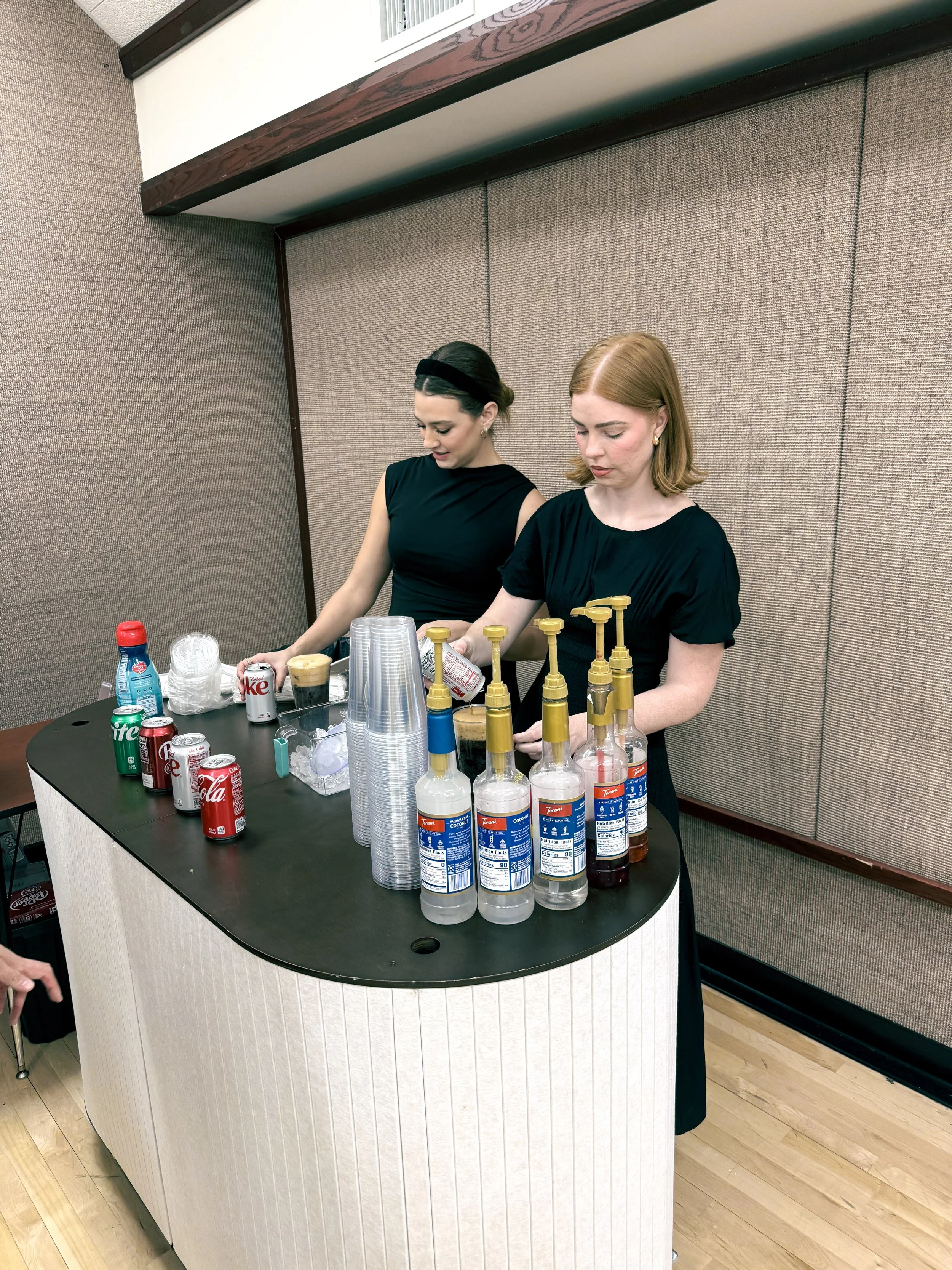 Two women in black dresses standing behind a drink station with soda cans, glass bottles with syrup pumps, and plastic cups.