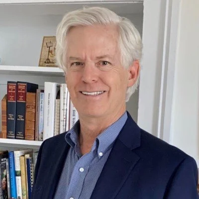 A smiling middle-aged man with white hair, wearing a blue collared shirt and dark blazer, standing in front of a bookshelf filled with books.