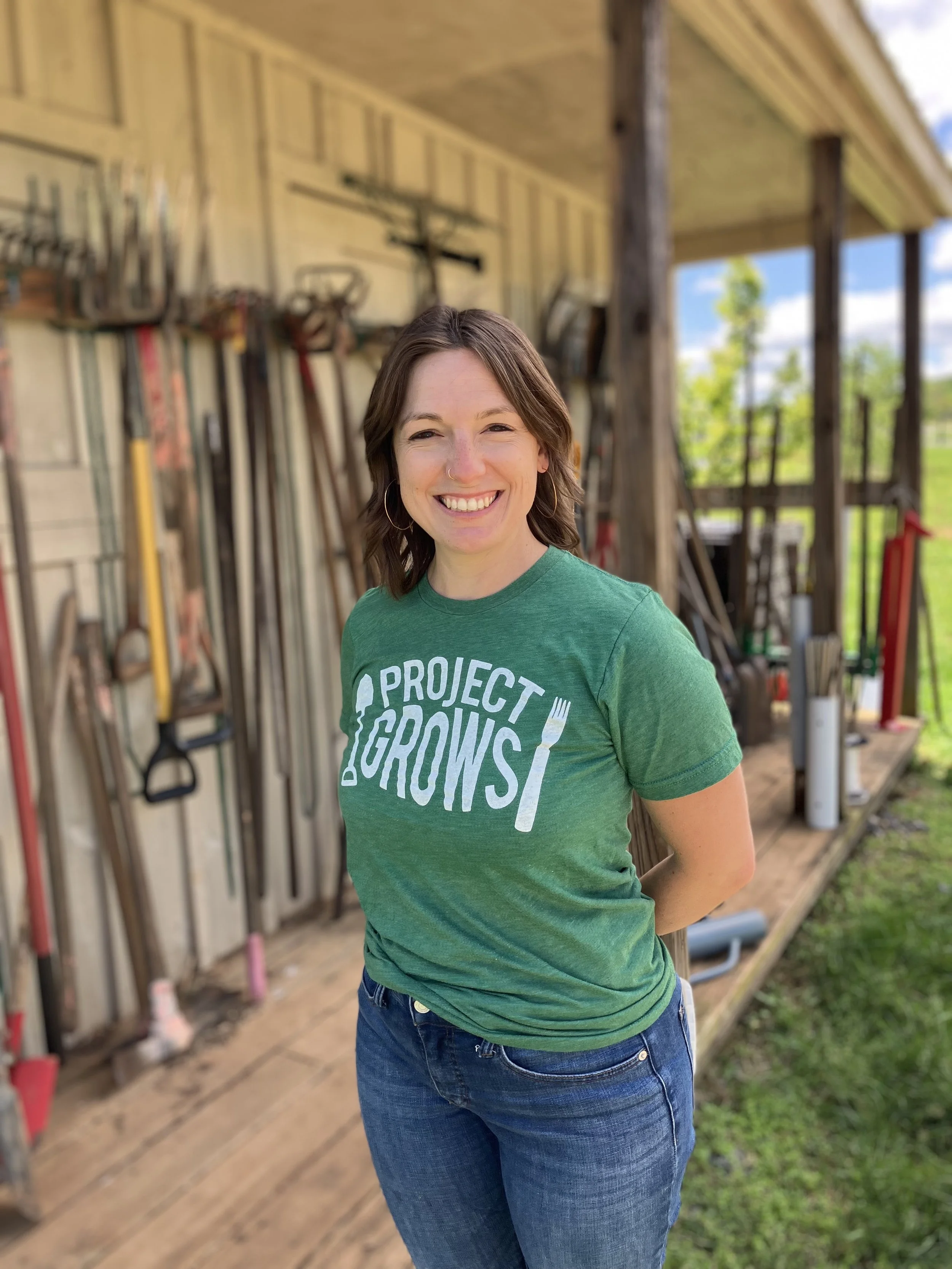 A woman with short brown hair, wearing a green t-shirt with "Project Grows" logo, smiling in a woodworking shed with tools hanging on the wall behind her.