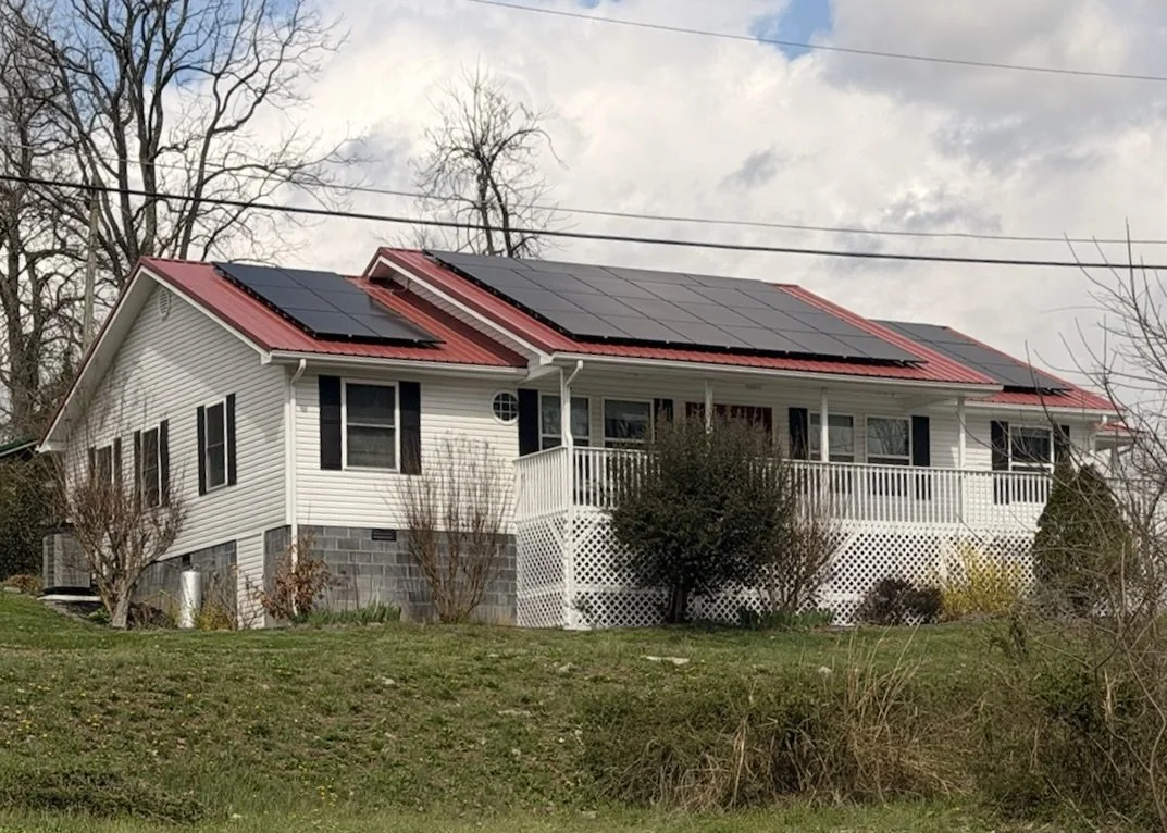 Single-family house with white siding and a red metal roof equipped with black solar panels, green front yard, trees, and cloudy sky.