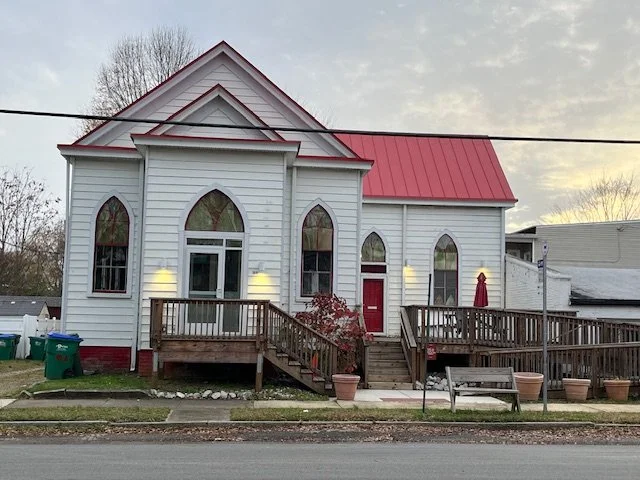 A white wooden church with a red roof and arched windows, surrounded by a wooden deck and some potted plants, set against a sunset sky.