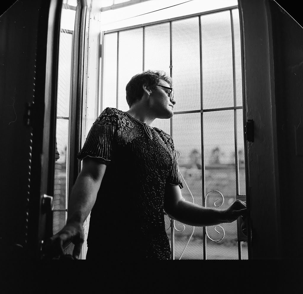 A man with short hair and glasses standing at a door with a screen, looking outside, in black and white.