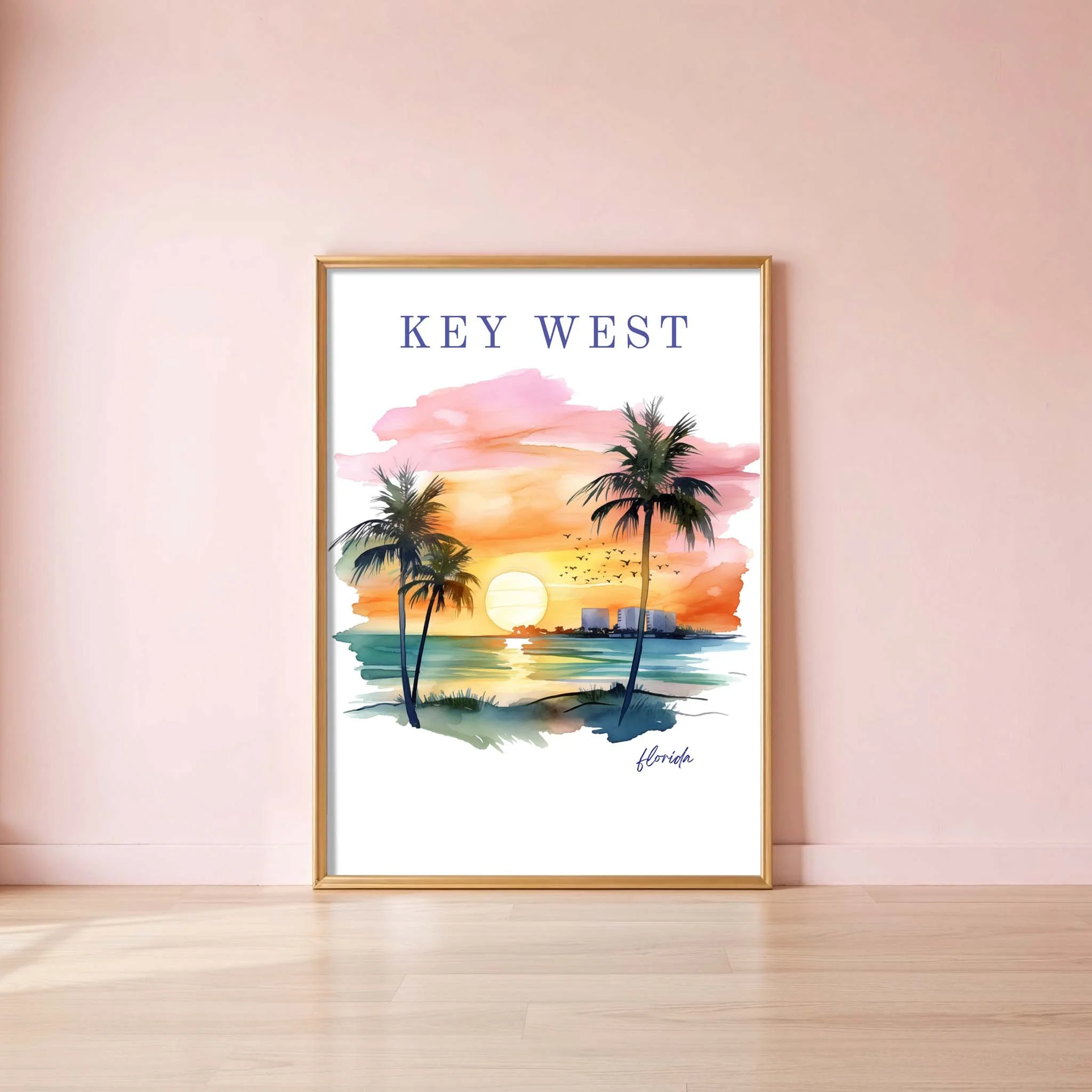 Framed watercolor print of a sunset over a beach with palm trees, titled "Key West" and labeled "Florida" in the bottom right corner, leaning against a pink wall on a light wood floor.