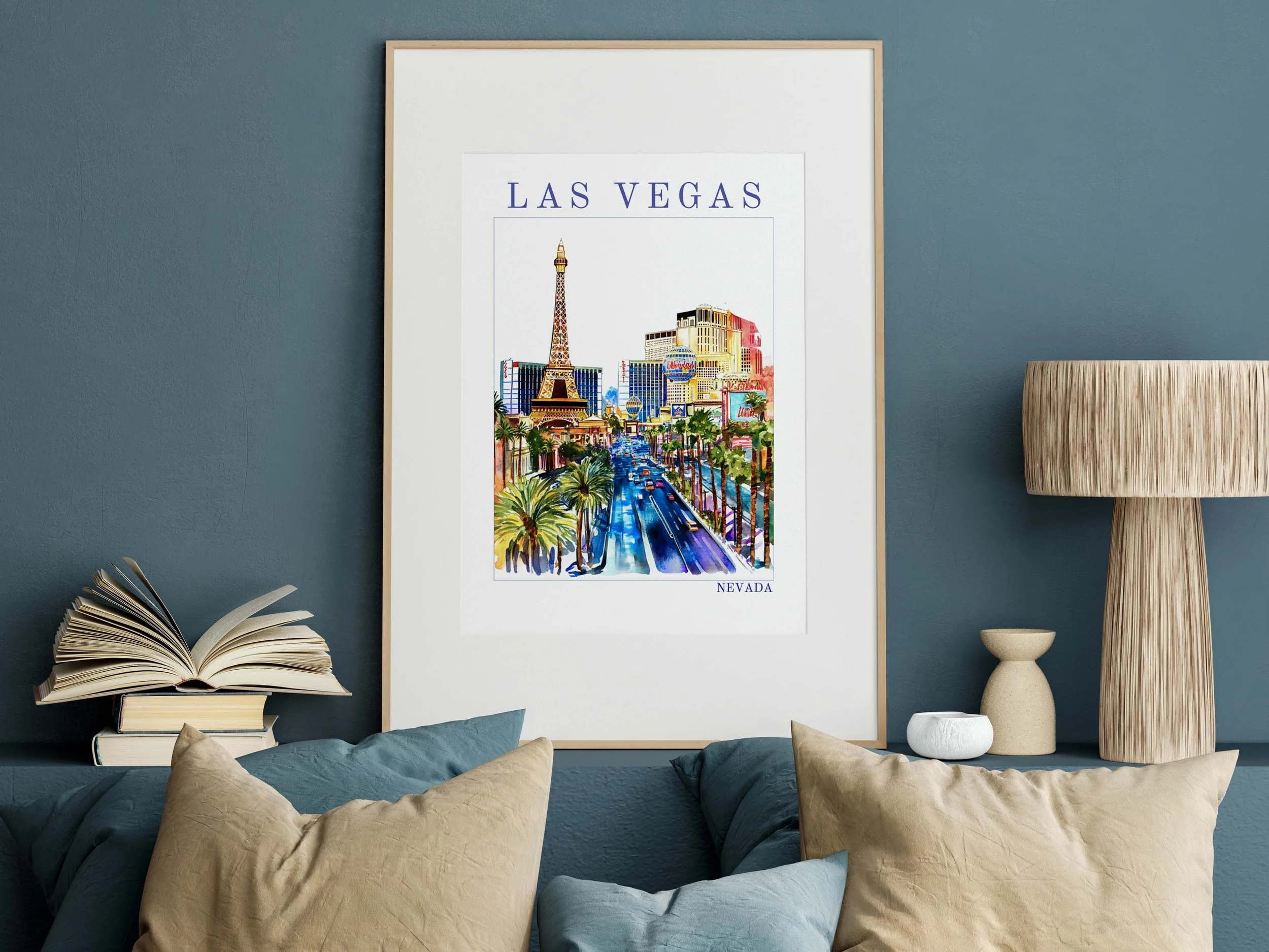Colorful watercolor poster of Las Vegas with the Eiffel Tower replica, high-rise buildings, palm trees, and a busy street, framed and hanging on a dark teal wall, next to a standing lamp and decorative vases, with pillows on a sofa below.