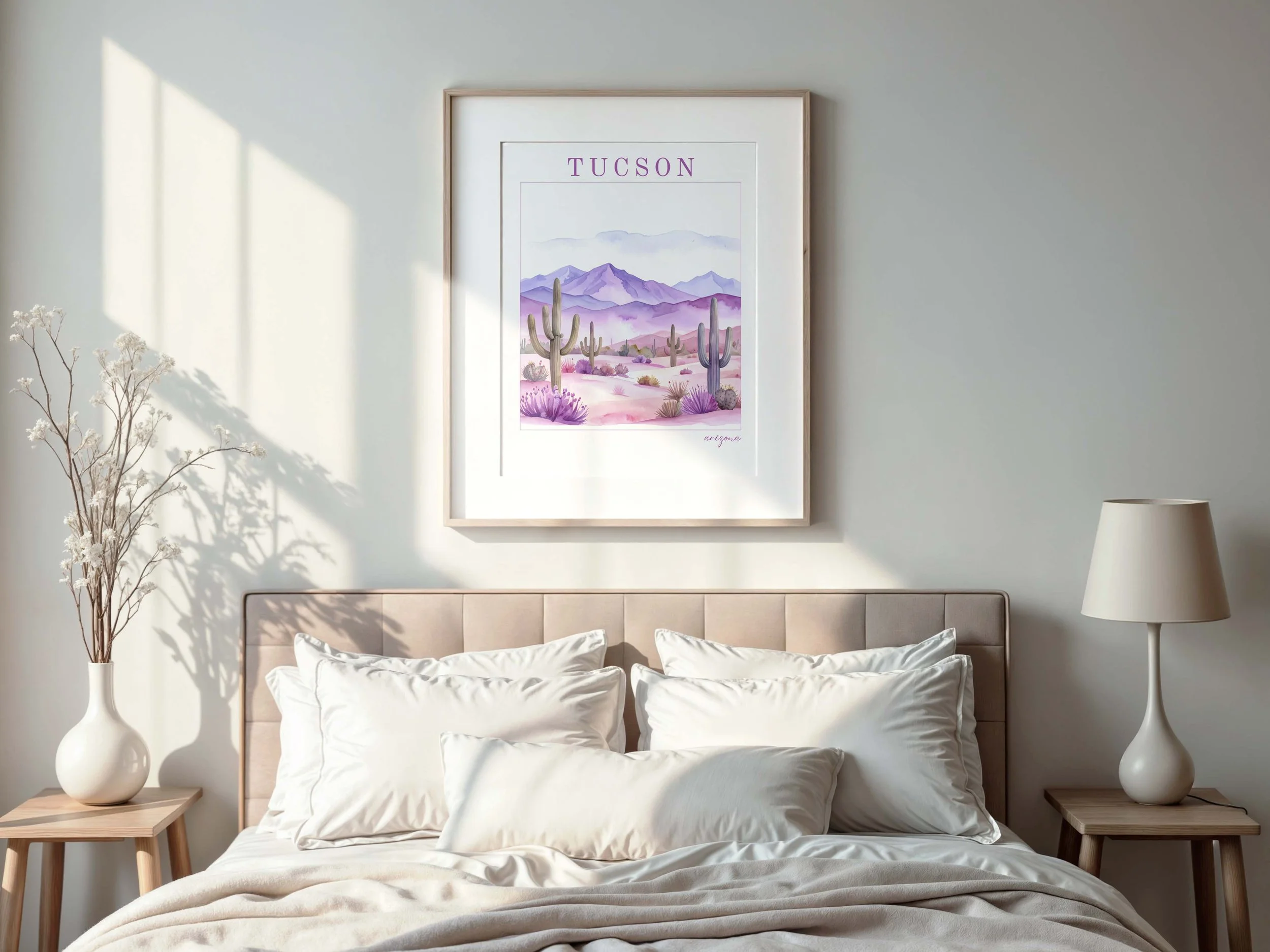A bedroom with a beige upholstered headboard, white bedding, and a wooden nightstand on each side. The left nightstand has a white vase with dried branches, and the right has a white table lamp with a beige lampshade. Above the bed hangs a framed art print of a desert scene with purple mountains, cacti, and the word 'TUCSON' at the top.