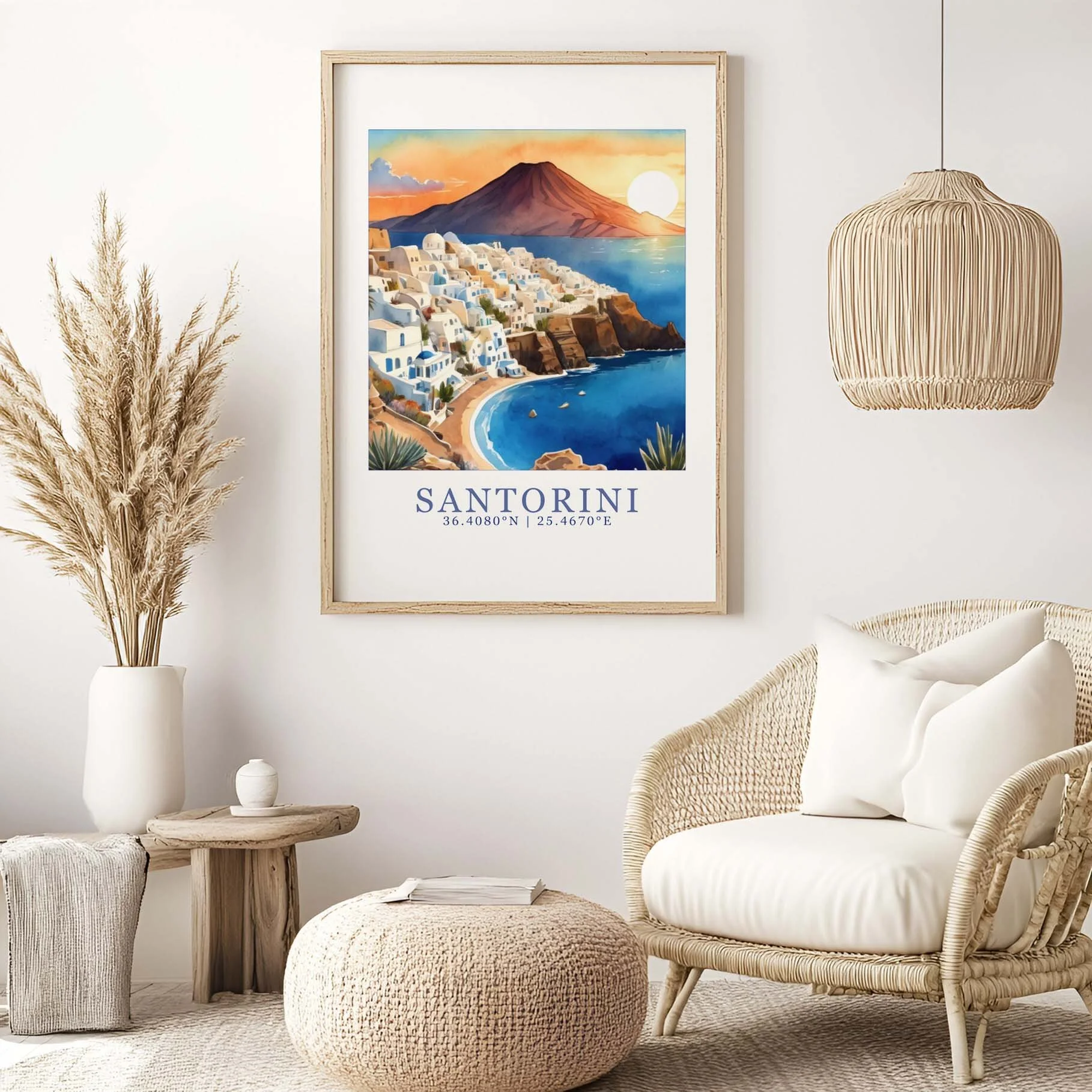 A living room corner with a framed poster of Santorini, Greece, featuring a volcano, white buildings, and a sunset, as well as a wicker chair with white cushions, a round woven ottoman, a small wooden side table with a white vase, and dried pampas grass in a white vase.