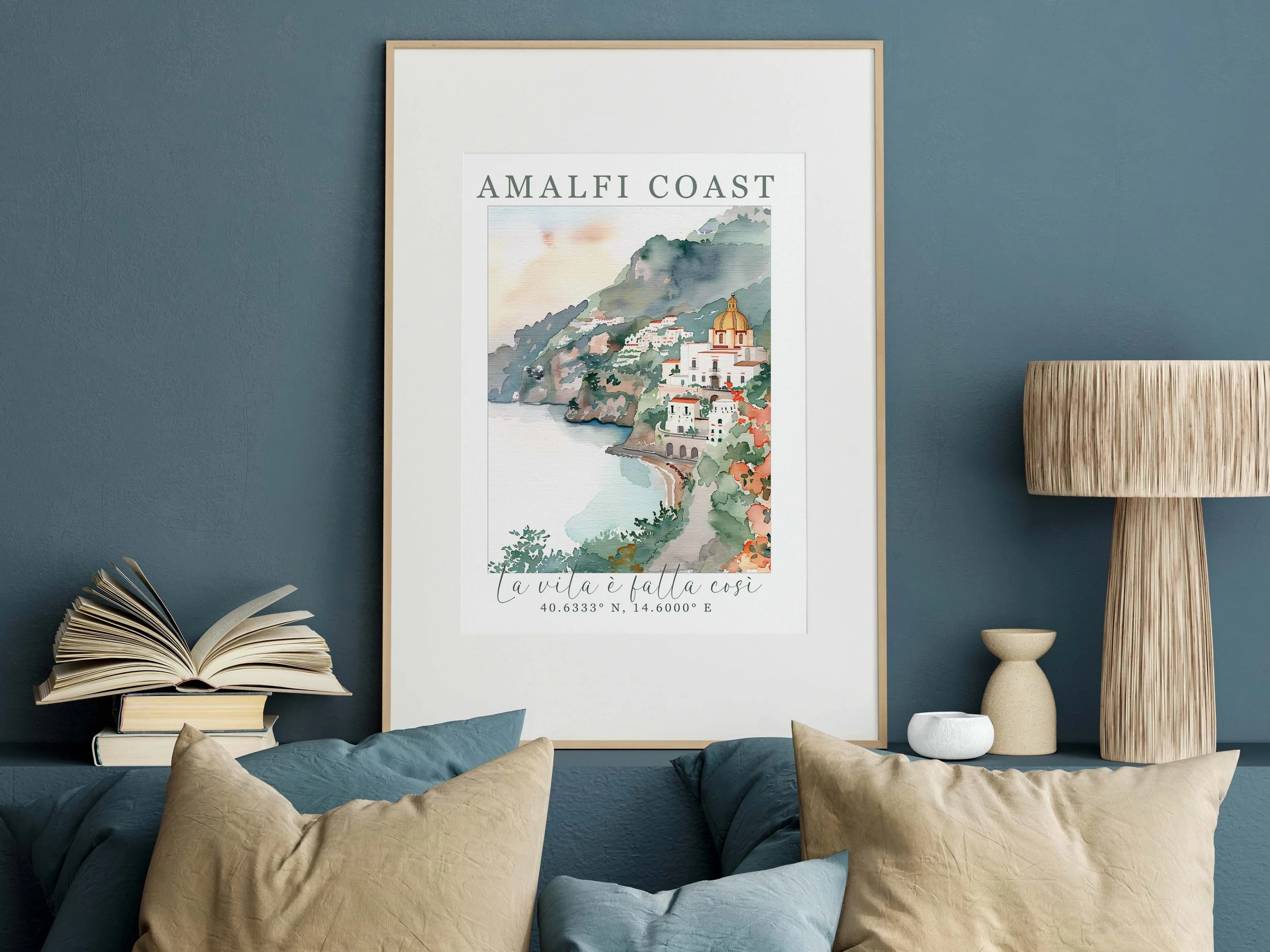 Decorative wall art poster featuring watercolor illustration of Amalfi Coast, with the text 'AMALFI COAST' at the top and a quote, 'la vila é fatta così,' along with geographical coordinates, with a table lamp, books, and cushions in a modern living room.