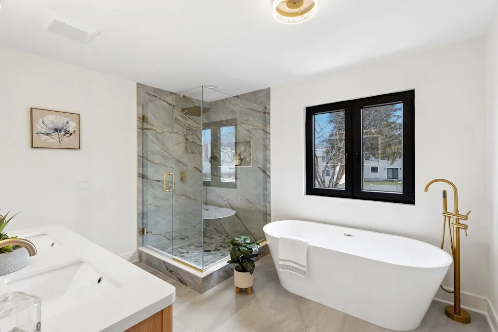 Modern bathroom with white freestanding bathtub, glass-enclosed shower with marble tiles, window with black frame, and gold fixtures.