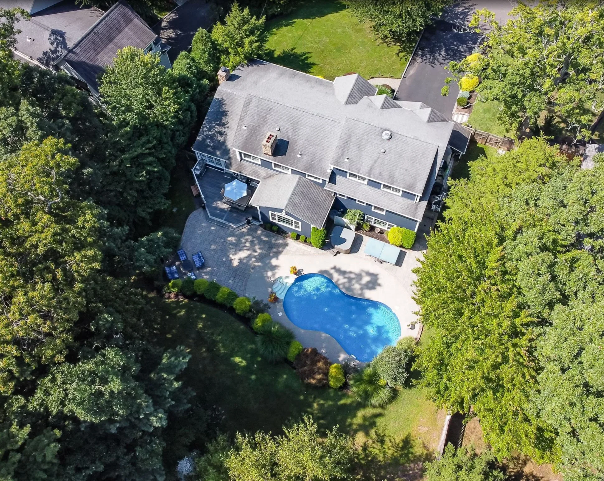 Aerial view of a backyard with a kidney-shaped swimming pool, surrounded by a patio with seating, lush green trees and grass, and a gray house with multiple roofs.