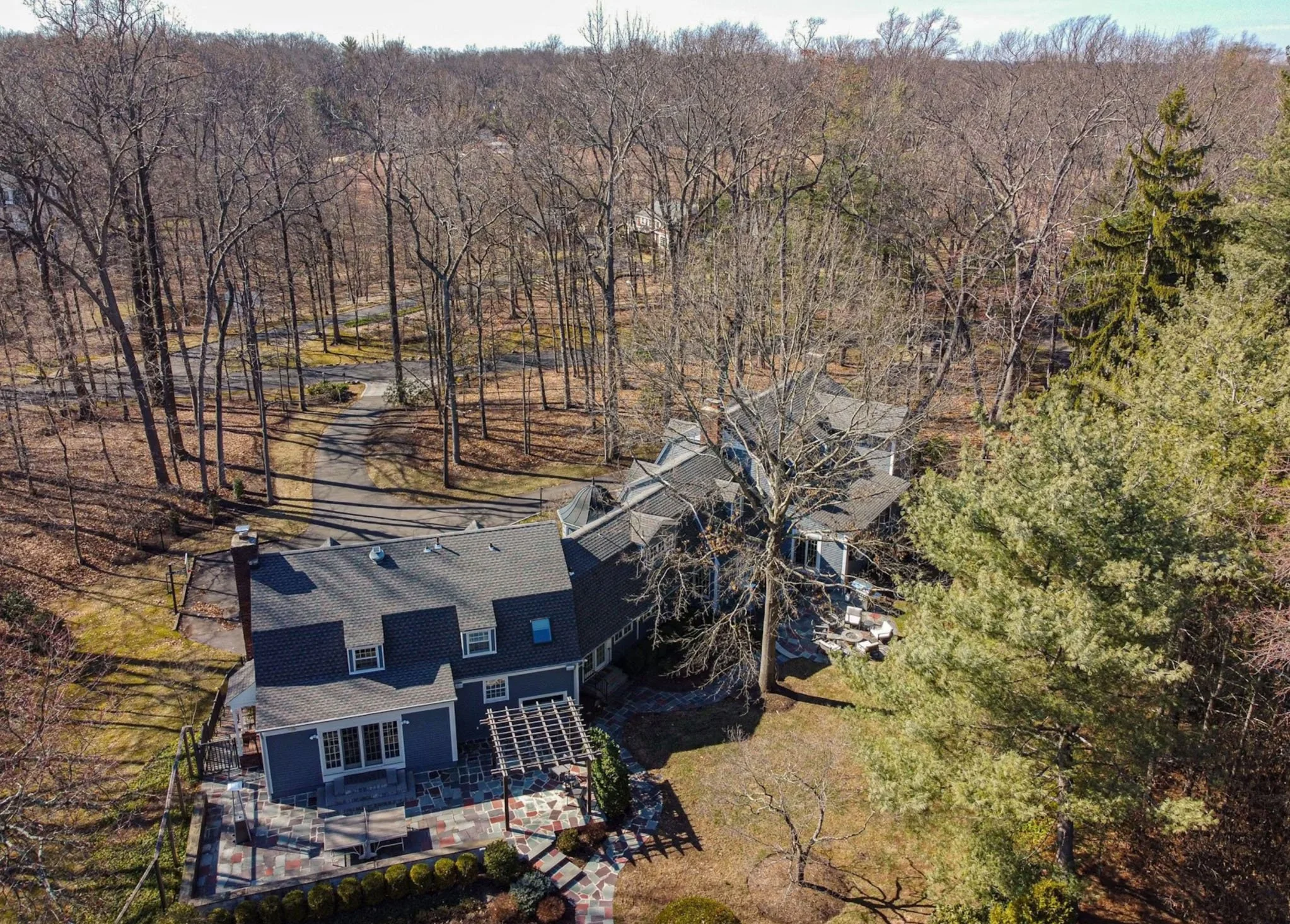 Aerial view of a house with a gray roof surrounded by trees and a winding pathway in a wooded area during late fall or early spring.