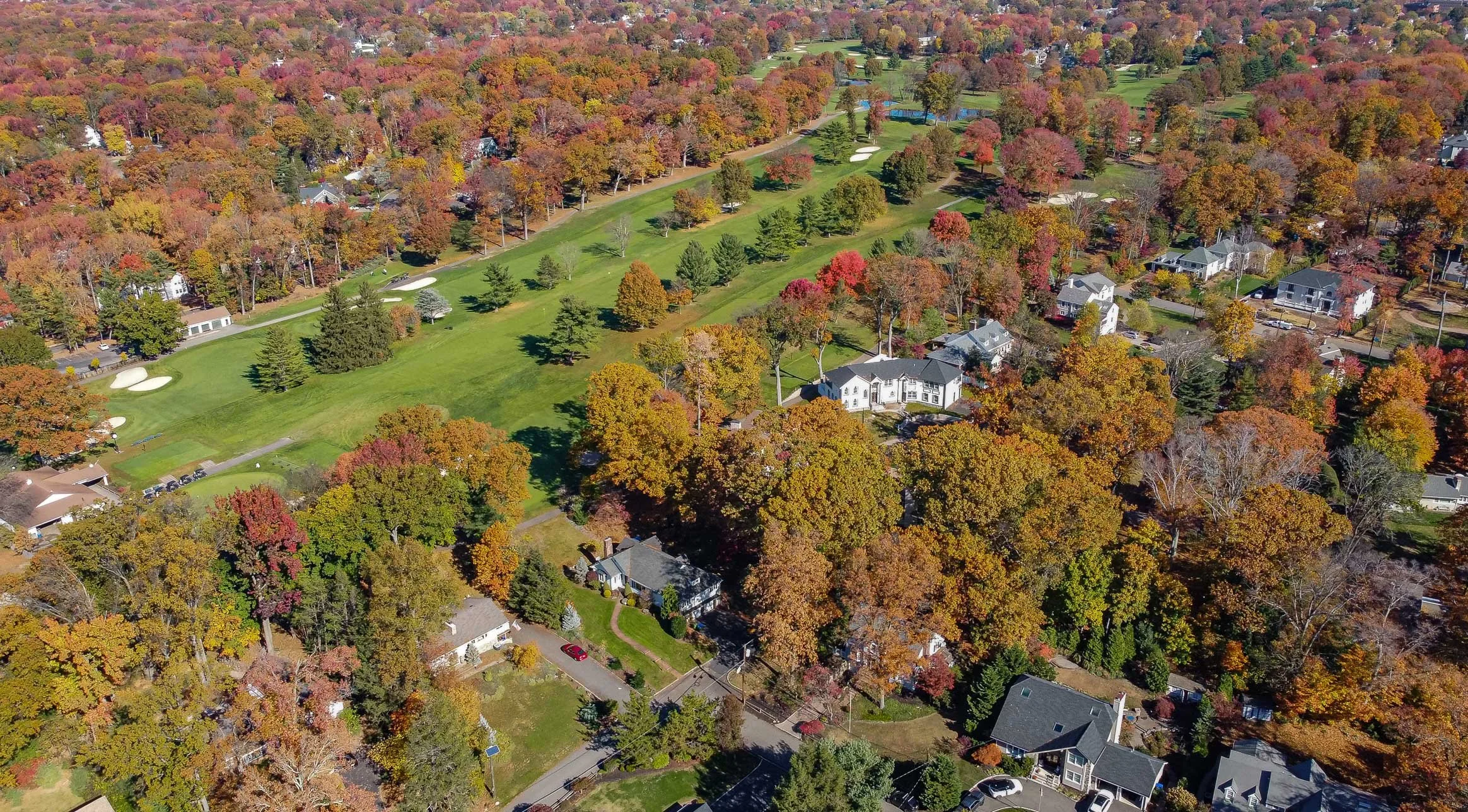 Aerial view of a suburban neighborhood with a golf course, paved roads, trees with fall foliage, and houses with yards.