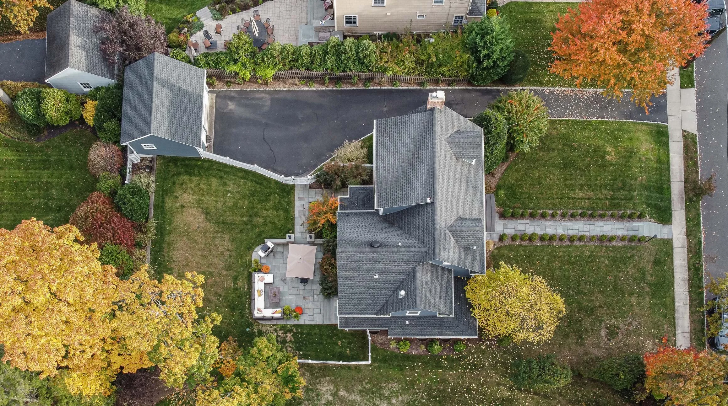 An aerial view of a house with a gray roof, surrounded by a well-maintained yard with green grass, trees with colorful fall leaves, a paved patio with outdoor furniture, and a driveway.