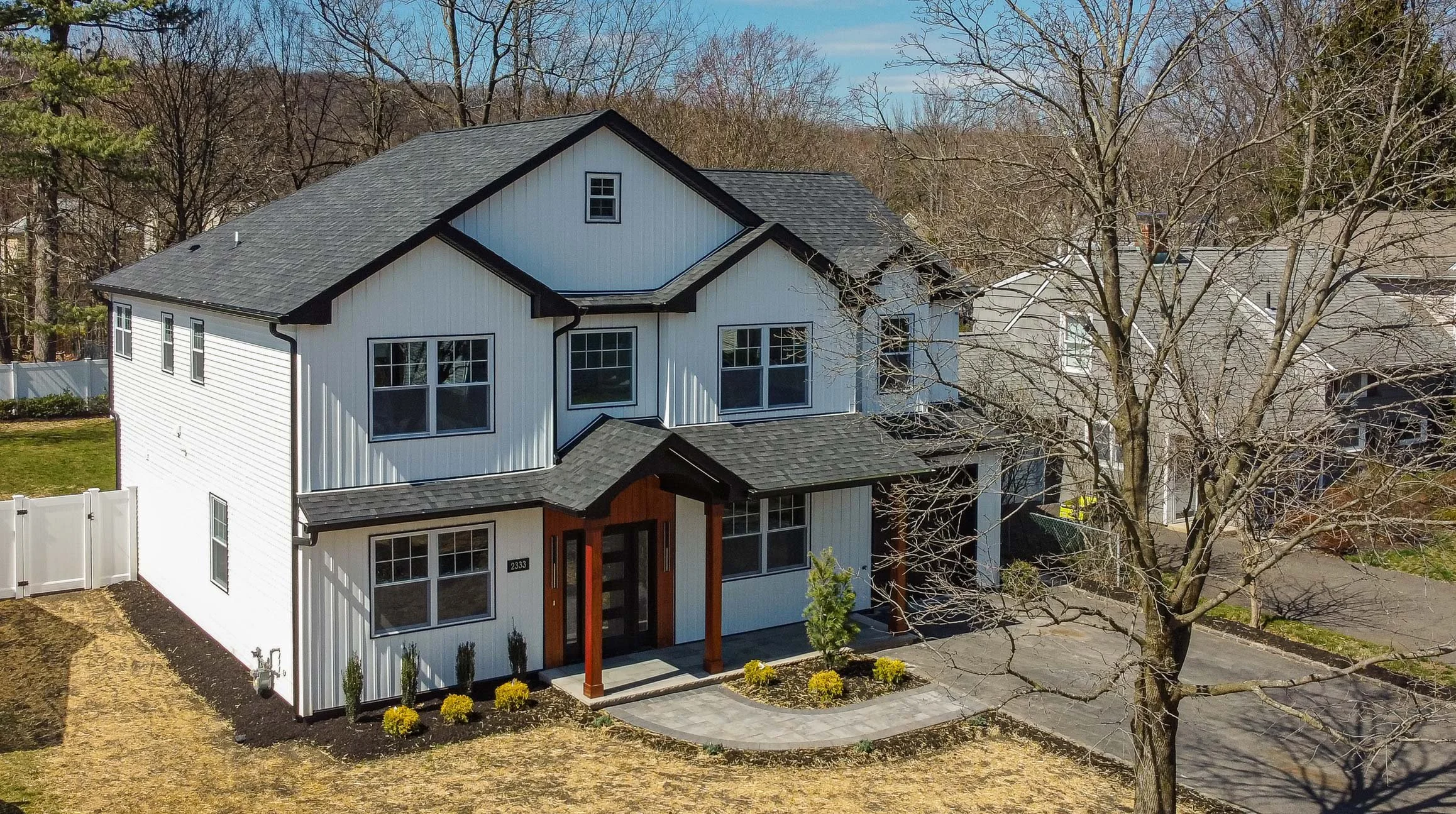 A two-story modern house with white siding, black trim, and a dark gray roof, surrounded by a yard with yellow flowers, a paved walkway, and leafless trees.