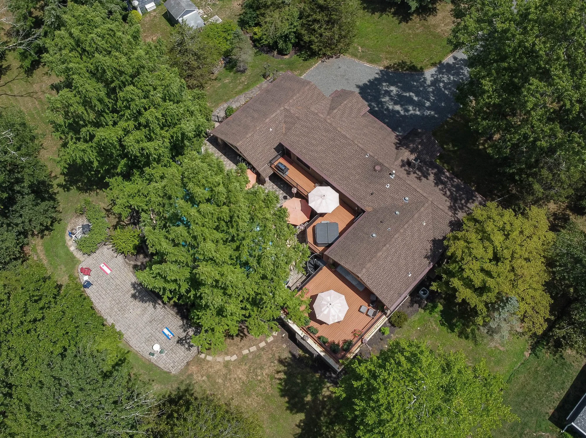 Aerial view of a house with a brown roof, surrounded by large green trees and a backyard patio with umbrellas and outdoor furniture.