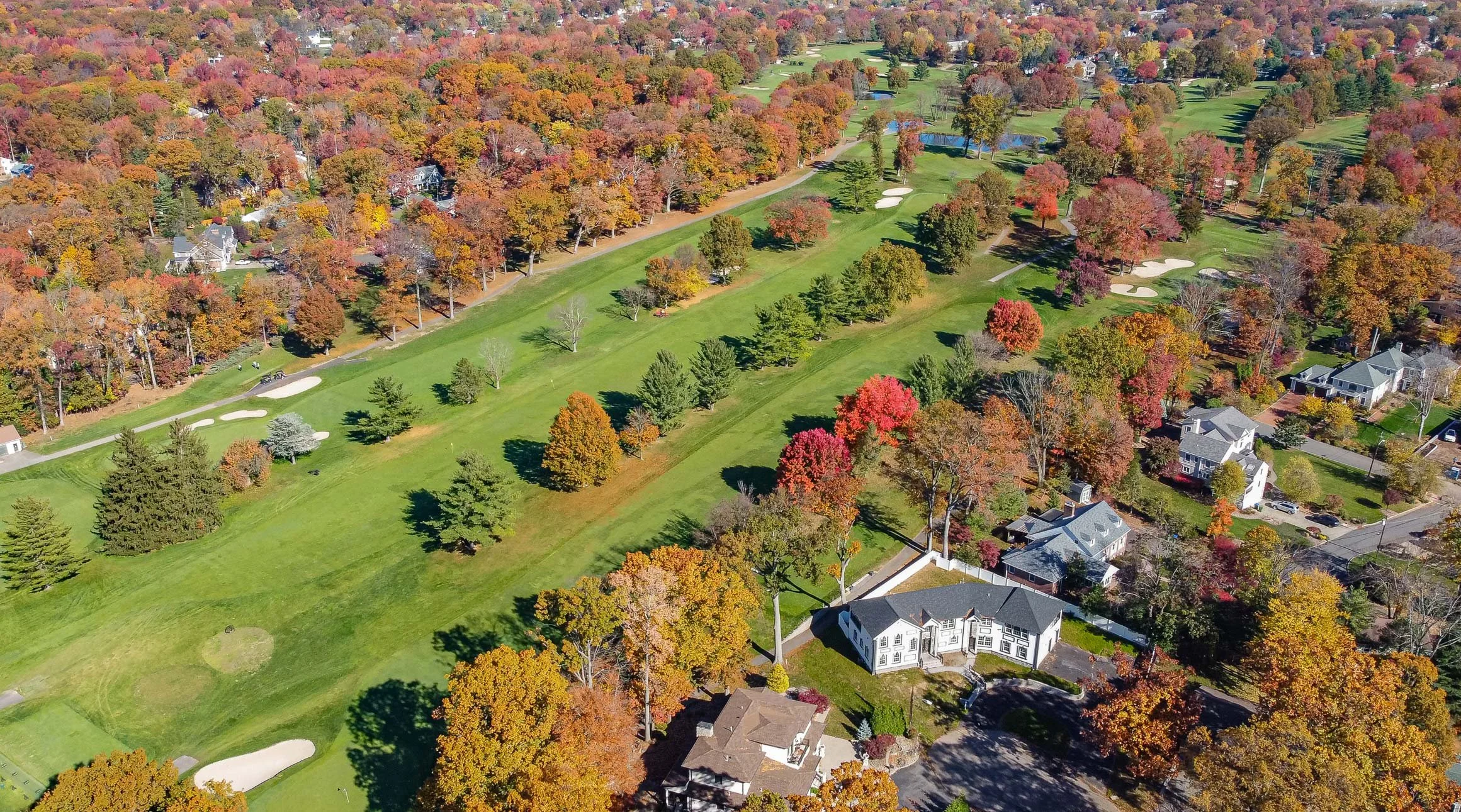 Aerial view of a colorful suburban neighborhood during autumn, showing a golf course, houses, and trees with vibrant fall foliage.