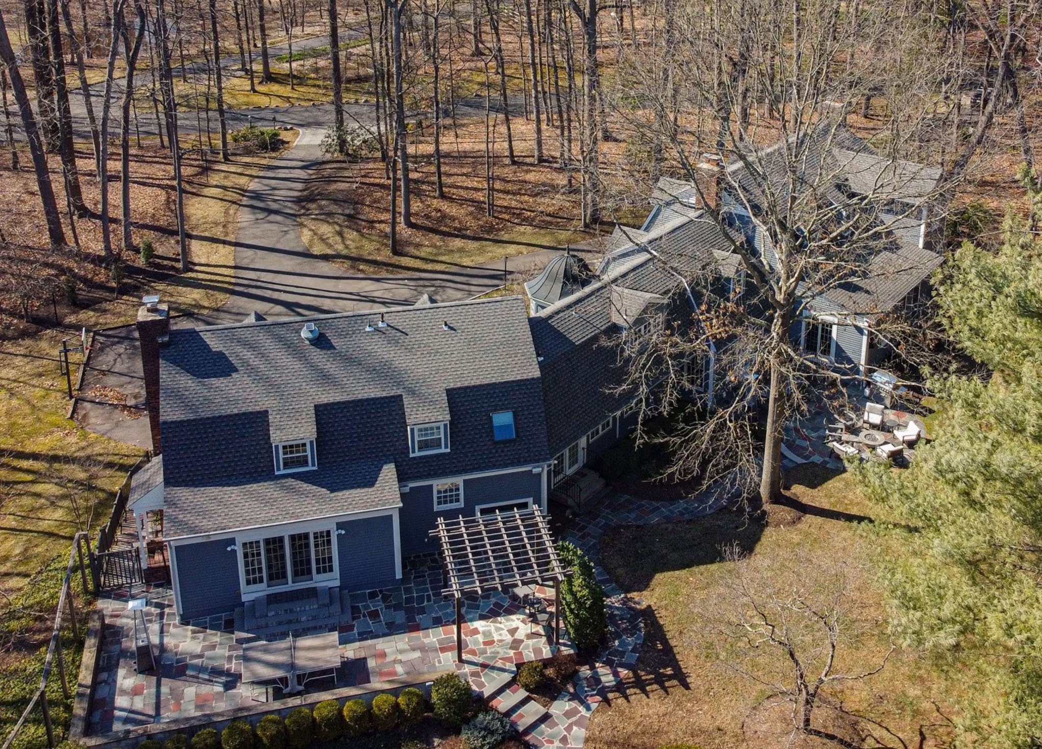 Aerial view of a two-story house with a gray roof, surrounded by leafless trees and a wooded backyard with a circular patio and outdoor furniture.