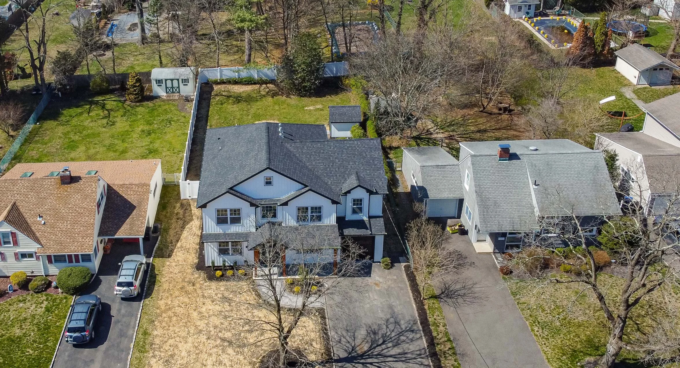 An aerial view of three houses in a neighborhood with lawns and trees, some with bare branches, and a fenced backyard with a garden and a shed.