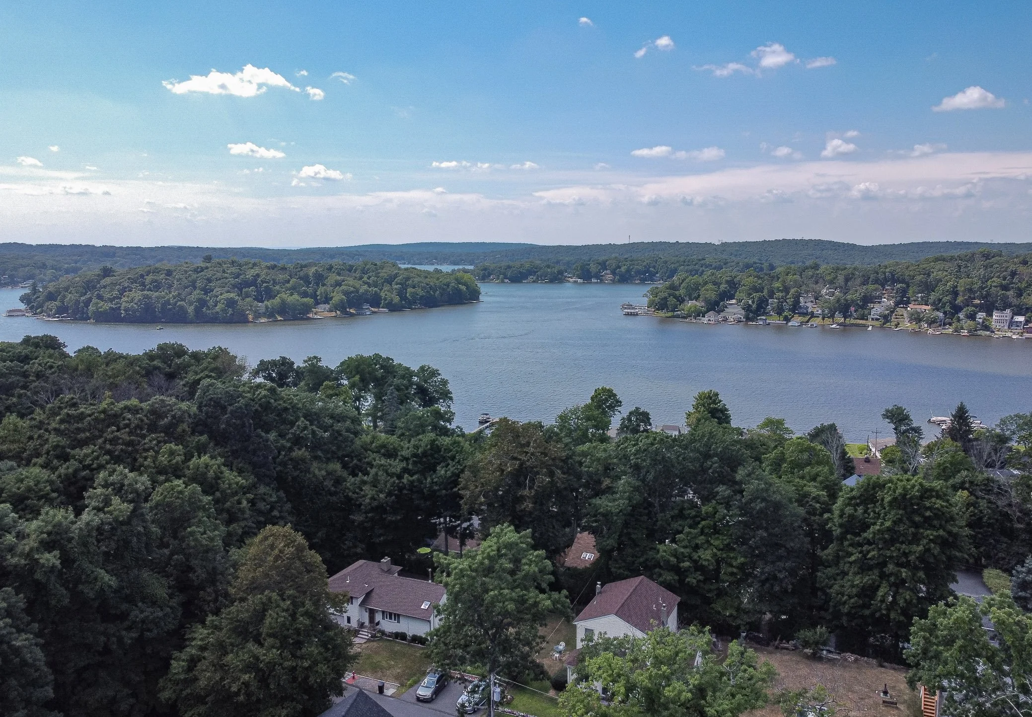 Aerial view of a lake surrounded by houses, trees, and hills under a partly cloudy sky.