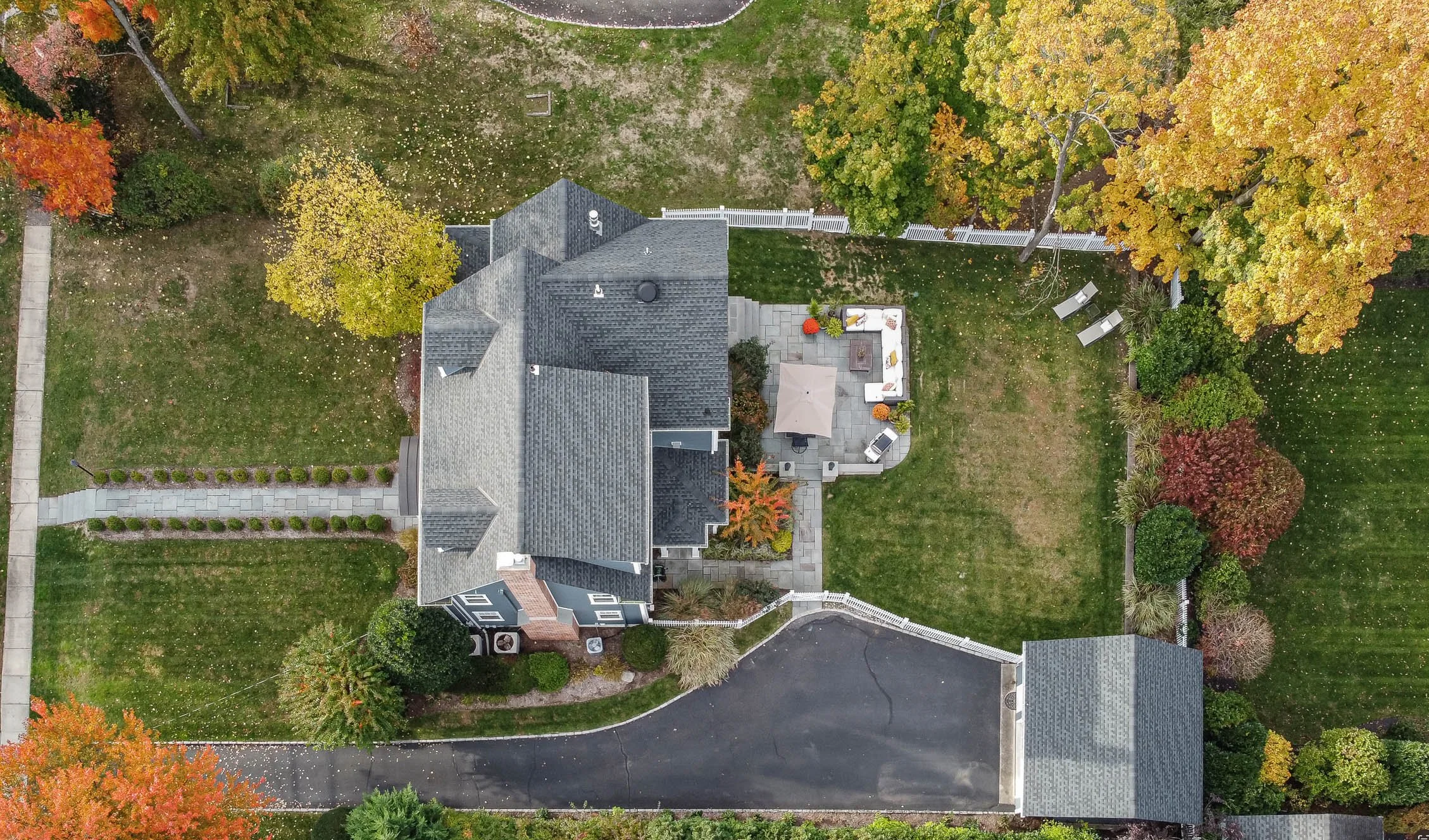 An aerial view of a house with a grey roof, surrounded by a backyard with trees showing autumn colors, a patio area with patio furniture, and a paved driveway.