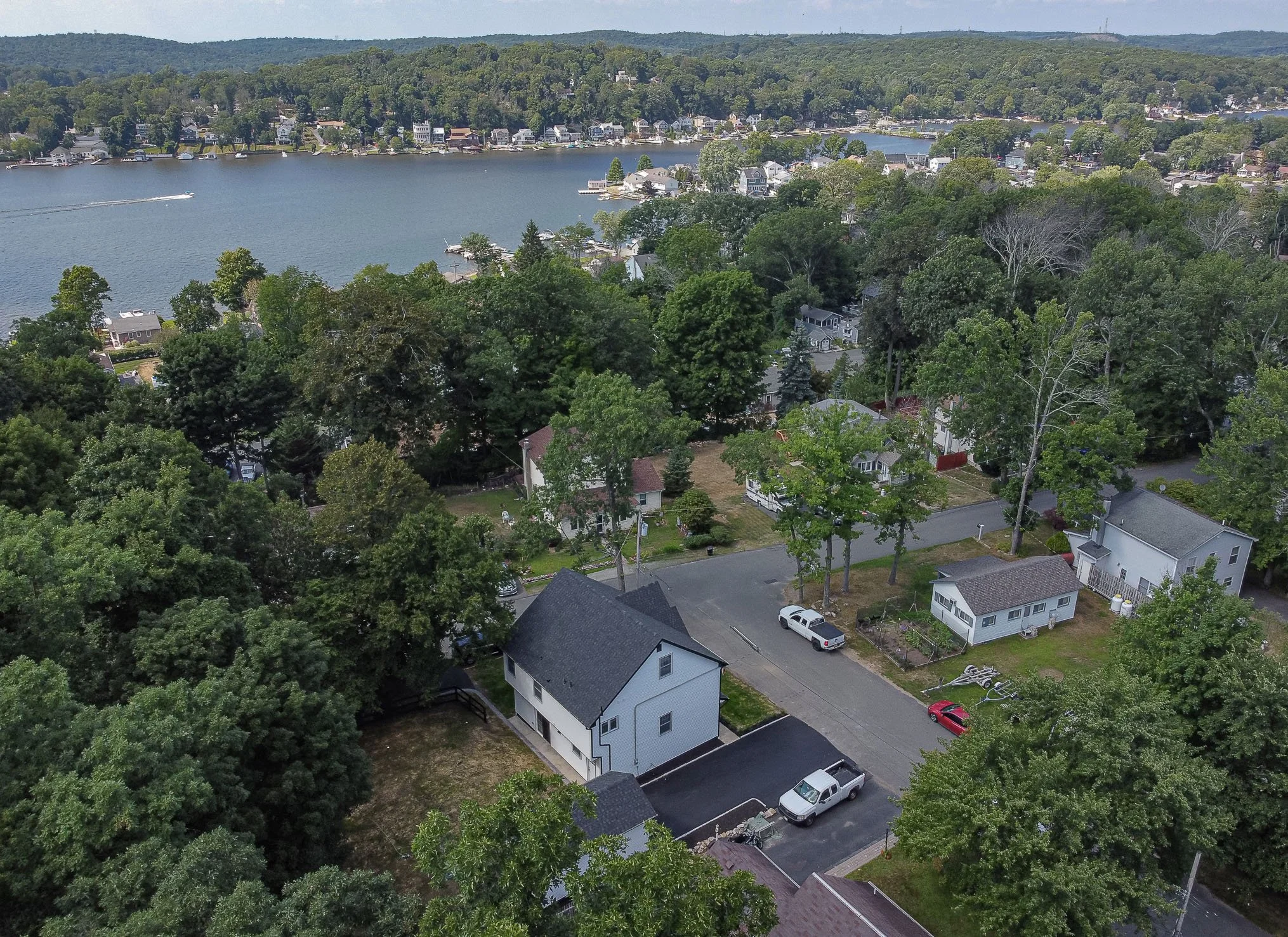 Aerial view of a lakeside suburban neighborhood with houses surrounded by lush green trees, and a large body of water with a boat in motion.