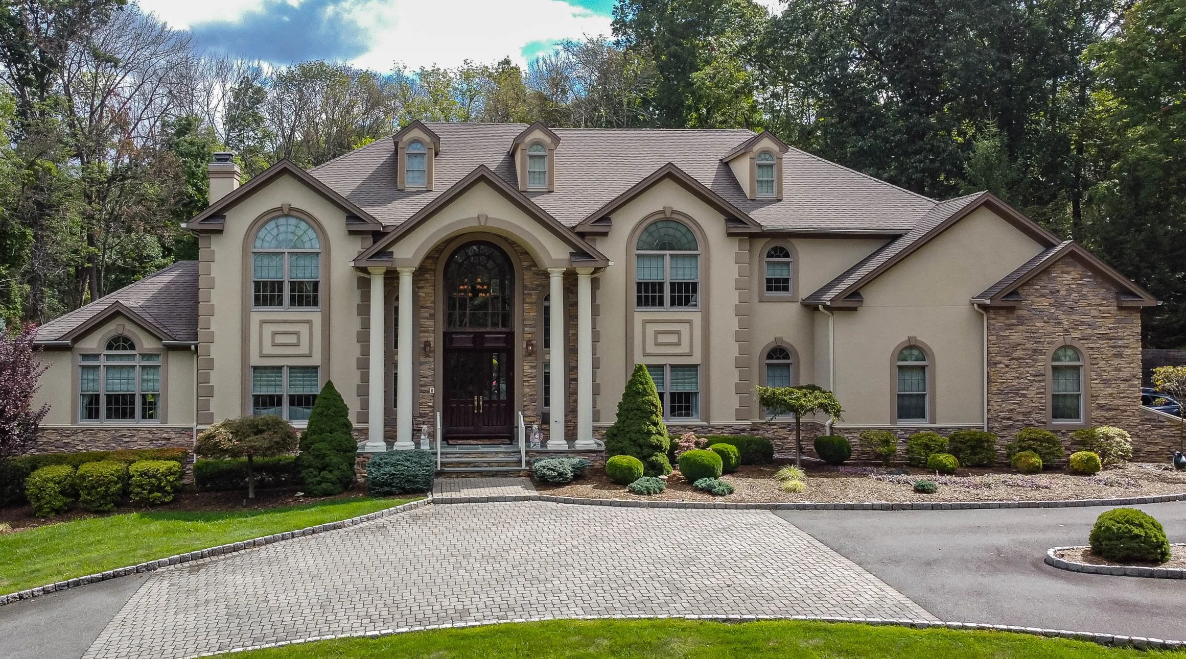 Front view of a large, two-story house with stone and stucco exterior, arched windows, a front porch with columns, and landscaped yard with shrubs and trees.