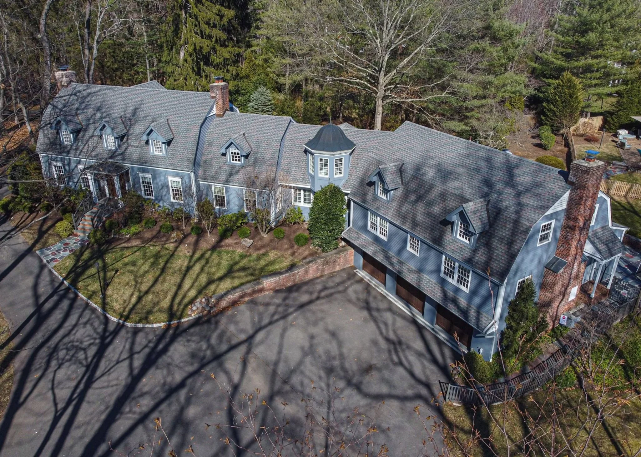 A large blue house with a gray shingled roof, multiple dormer windows, a brick chimney, a turret, surrounded by trees, with a driveway and garden in front.