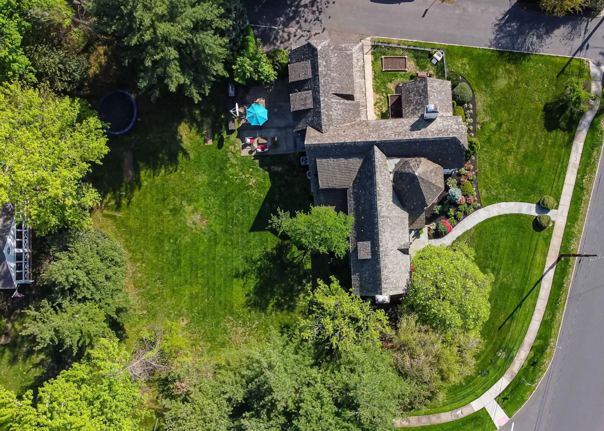 Aerial view of a house with a front walkway, surrounded by lush green lawn and trees, with a small backyard patio that includes outdoor furniture and a blue umbrella.