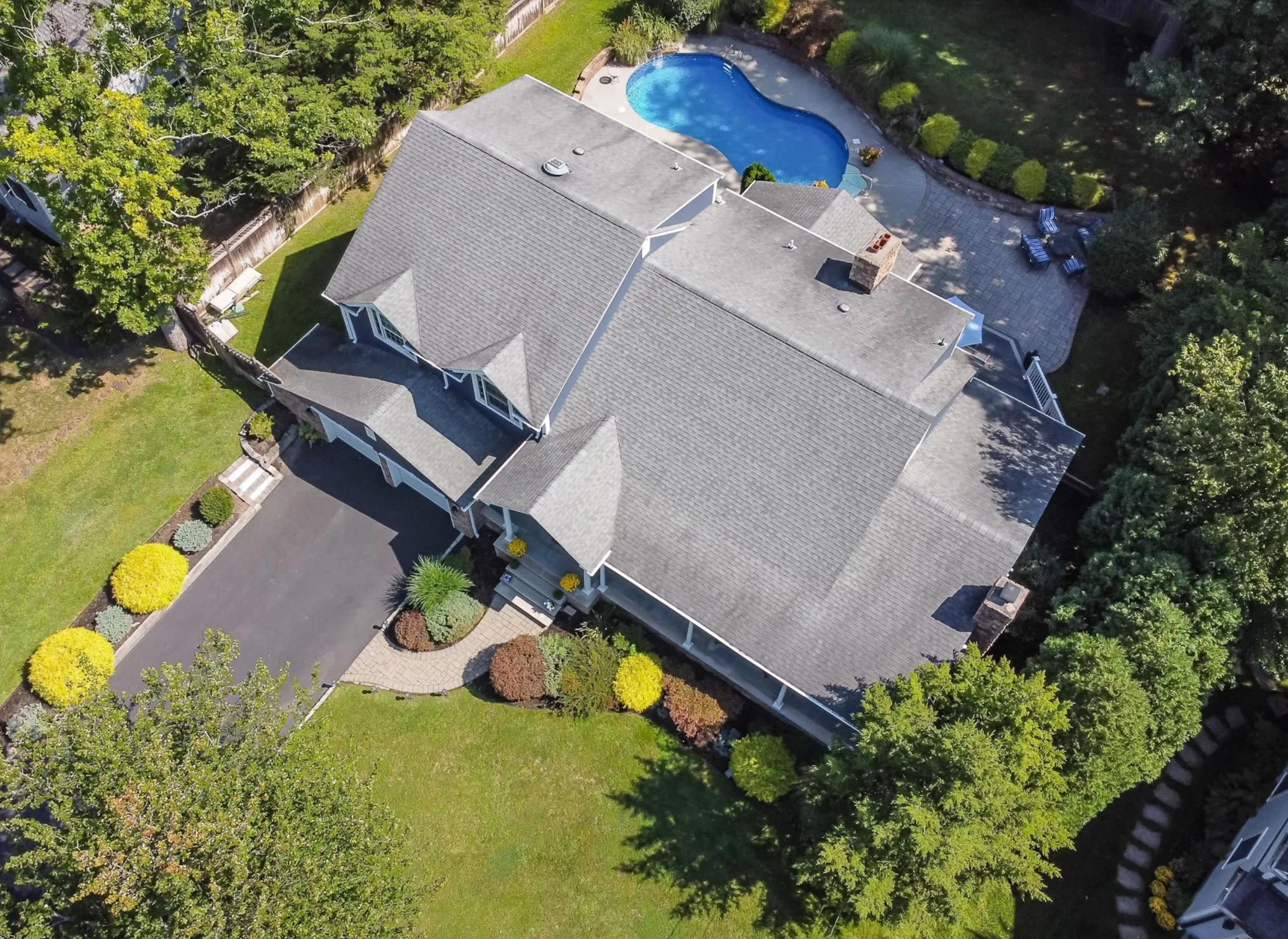 Aerial view of a house with a gray roof, surrounded by green trees and shrubs. The backyard features a kidney-shaped swimming pool, a paved patio area with outdoor seating, and a landscaped garden with various bushes and trees.
