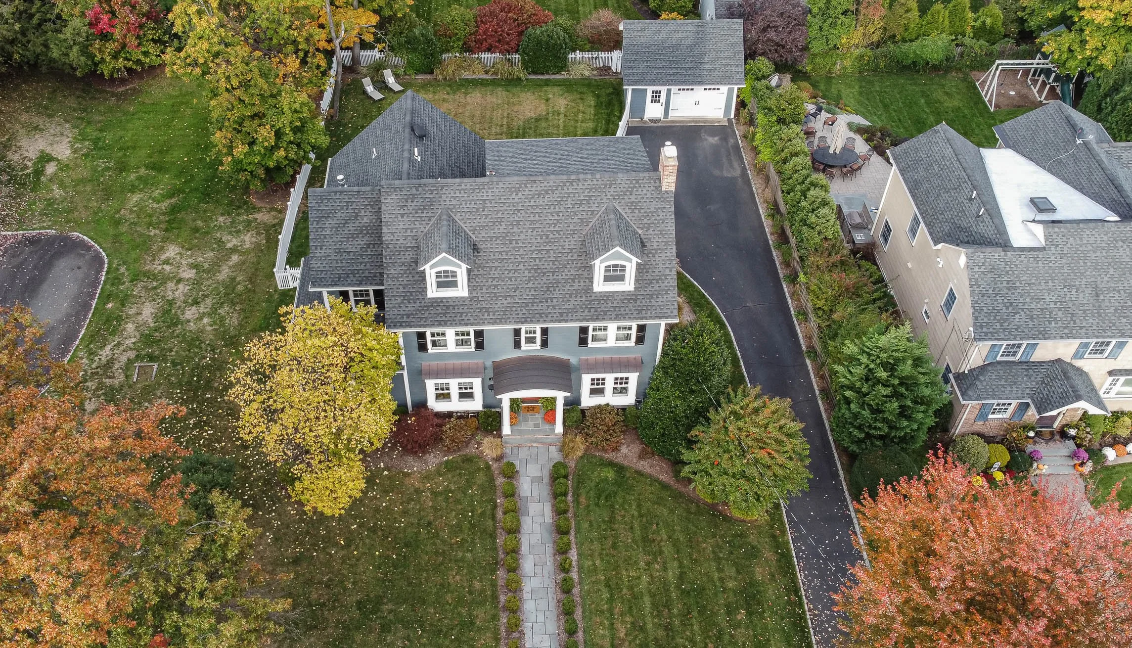 An aerial view of a house with a gray roof, surrounded by a lawn and trees with fall foliage. The house has a walkway leading to the front entrance and a driveway to a garage, with neighboring houses visible on the sides.