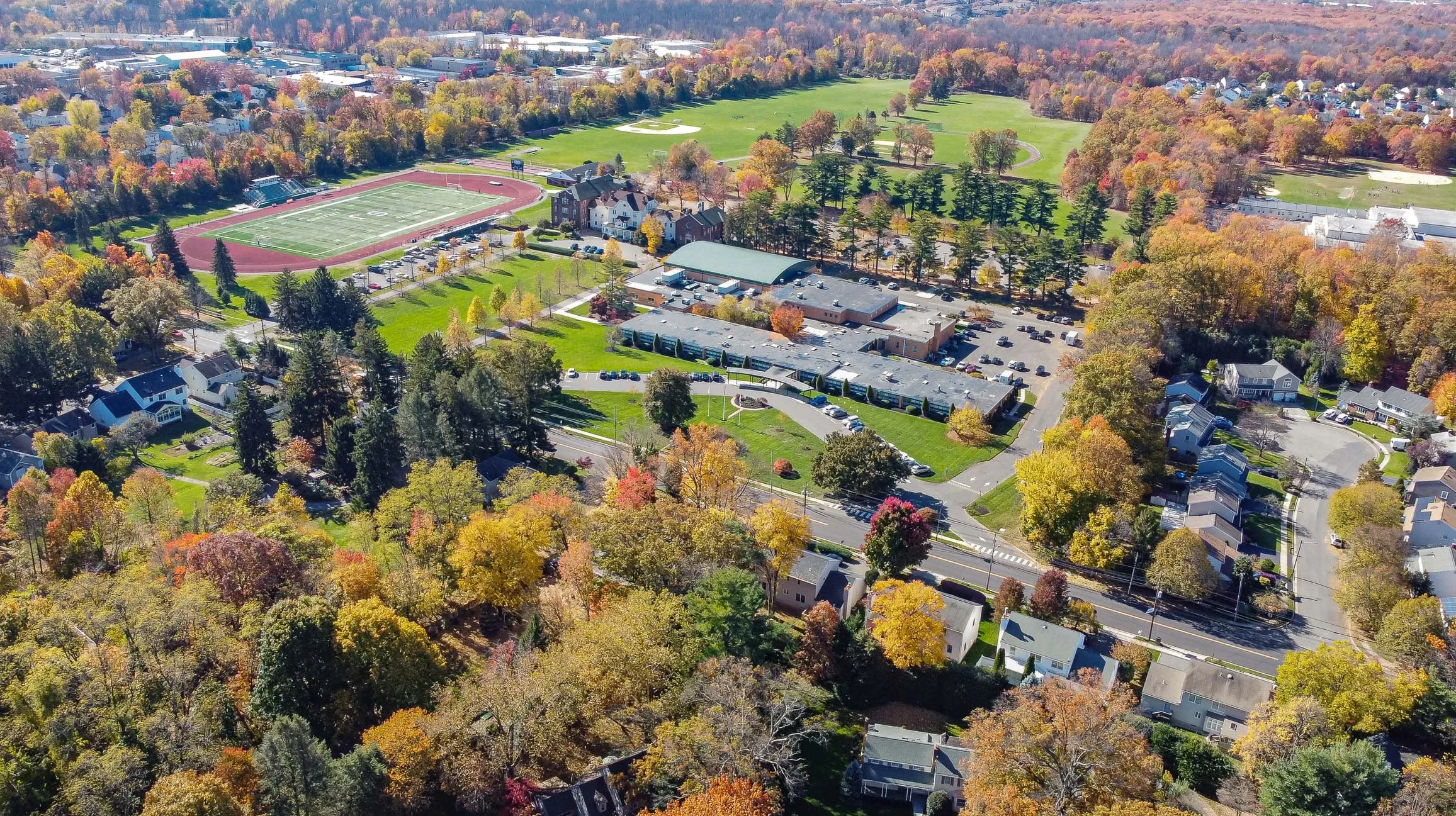 An aerial view of a suburban neighborhood surrounded by trees with fall foliage, a school with sports fields, houses, and parking lots.