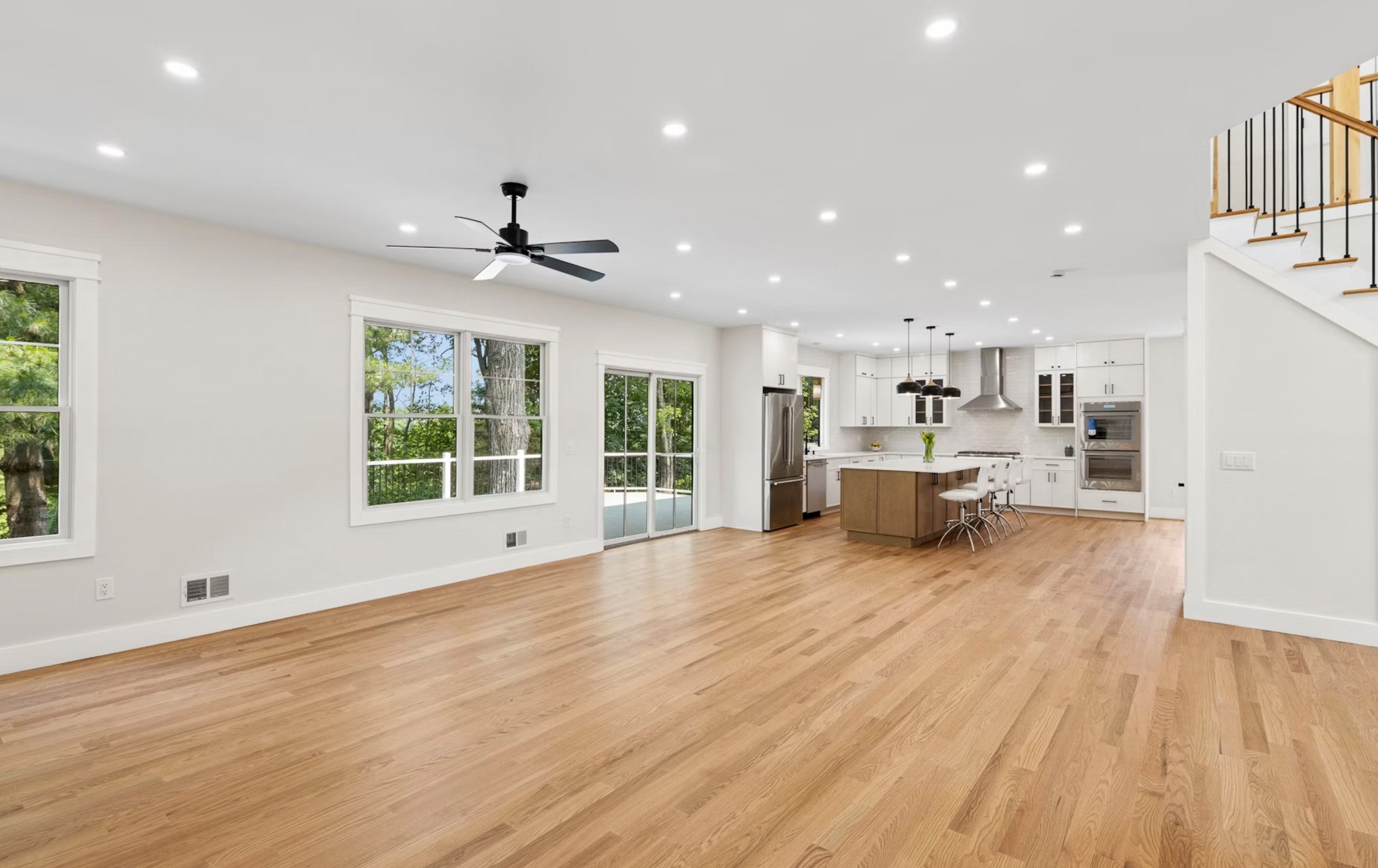 Open concept living room and kitchen with hardwood floors, white walls, large windows, and a staircase on the right side.