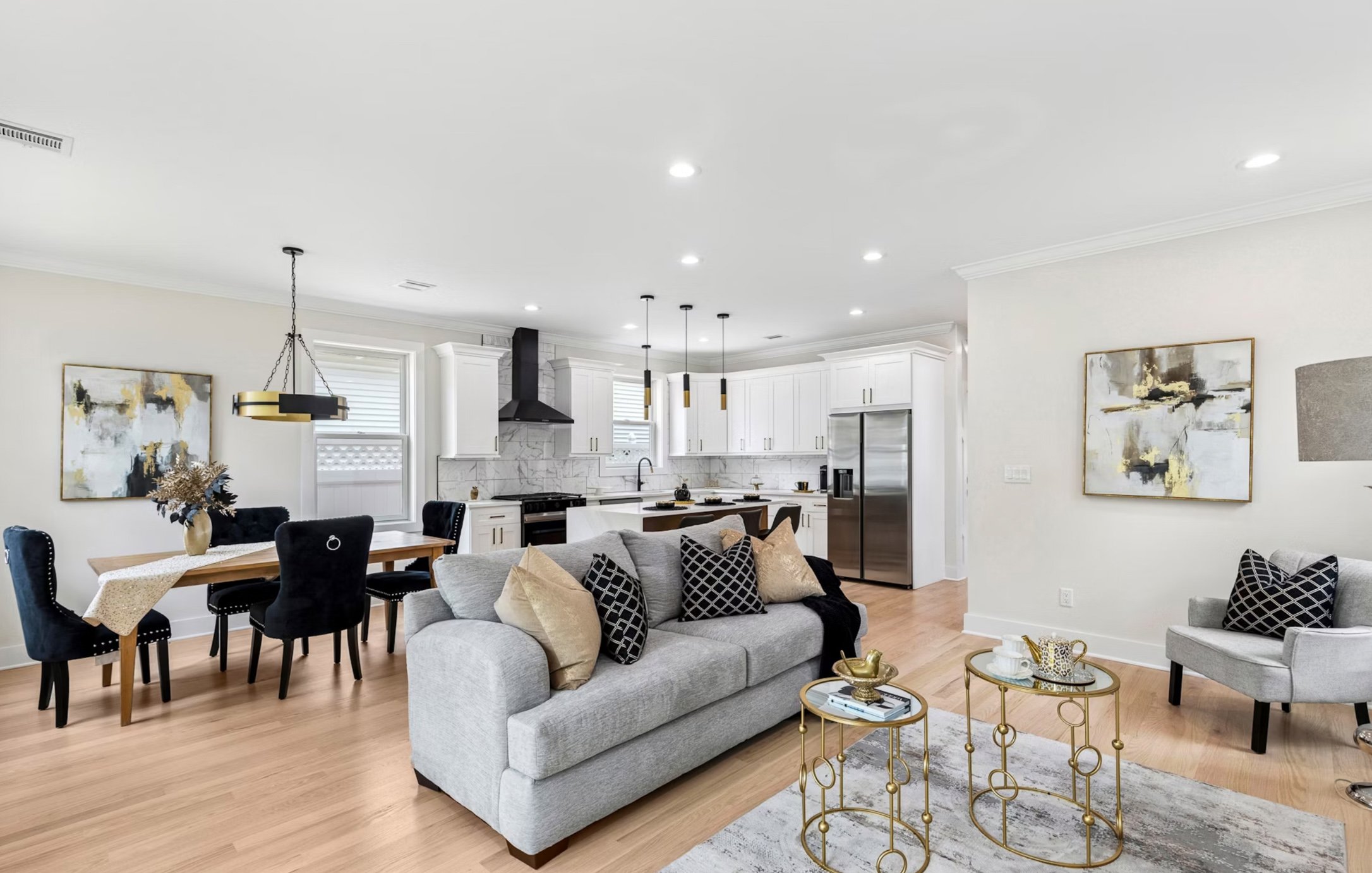 Open-concept living and dining area with a modern kitchen, light-colored walls, hardwood floors, a gray sofa with decorative pillows, a gray armchair, and gold accent tables, decorated with art and accessories.