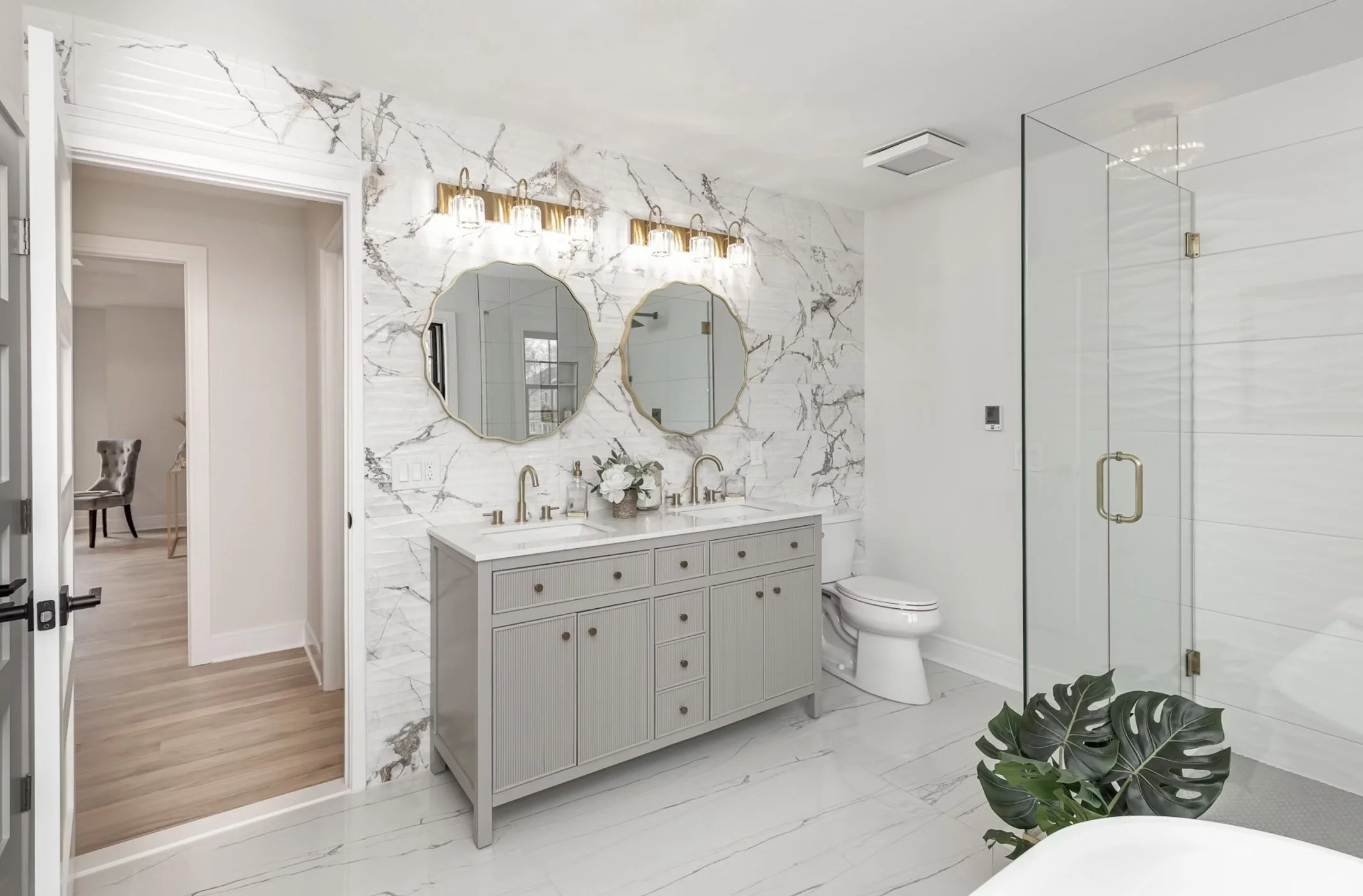 Modern bathroom with marble walls and flooring, double sink vanity with two mirrors, two light fixtures, and a glass shower enclosure. There is a plant near the bathtub and a doorway to an adjacent room.