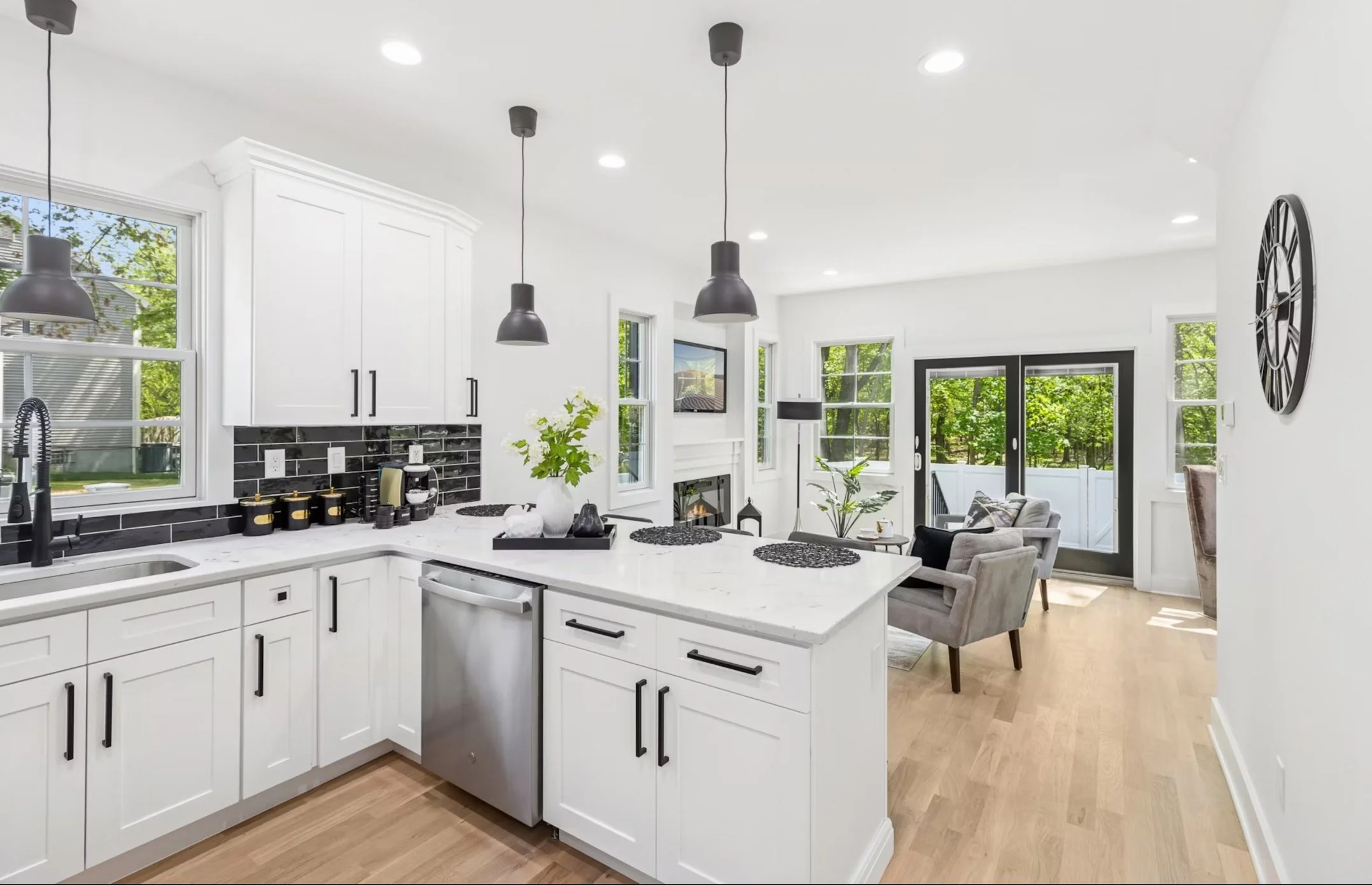 Modern kitchen with white cabinets, black hardware, black subway tile backsplash, and stainless steel appliances. Light wood flooring, black pendant lights, and a view into a bright living area with large windows and glass door.