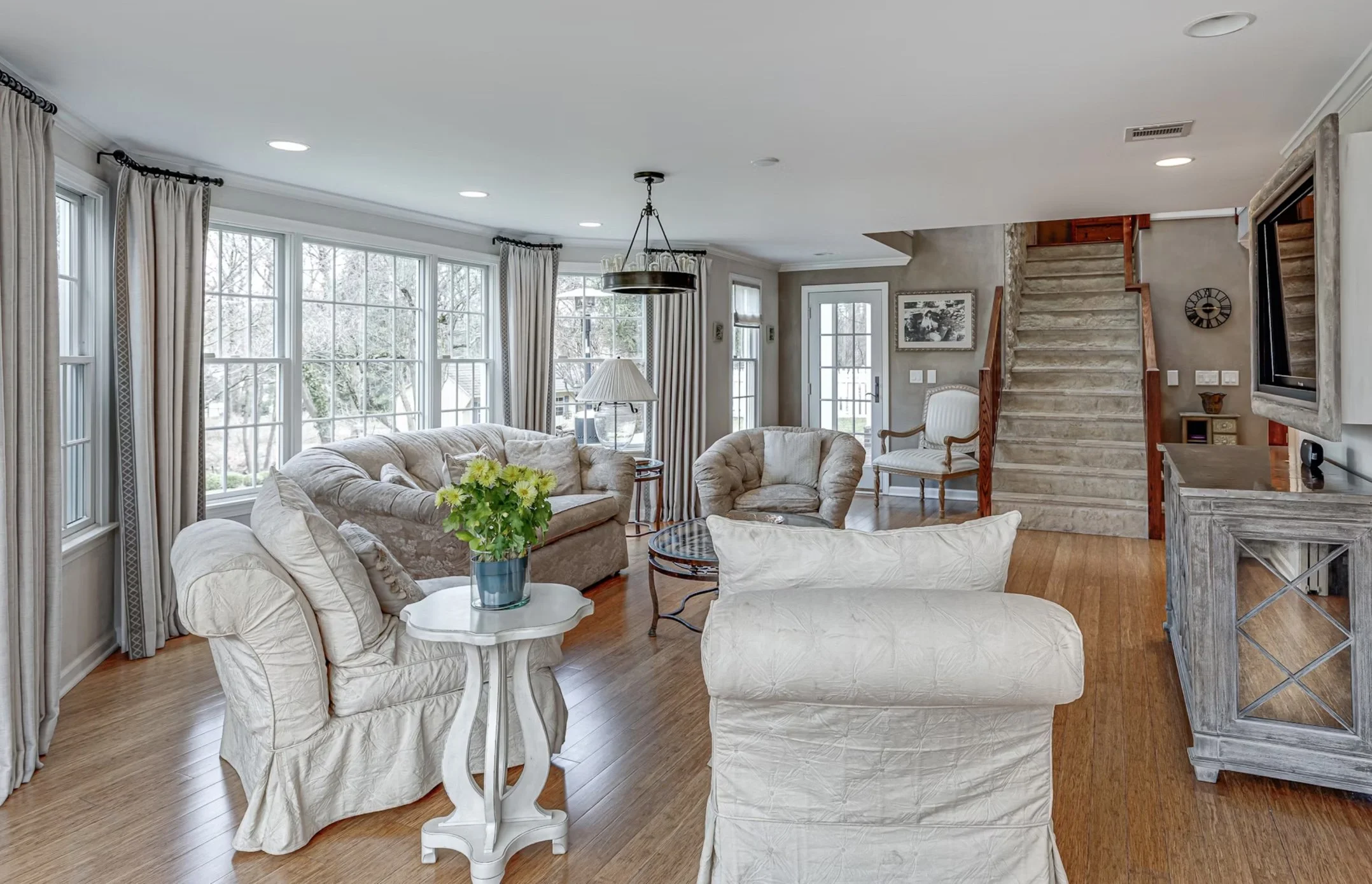 Bright living room with beige upholstered sofa and armchair, a small white side table with green flowers, large windows with curtains, hardwood floor, staircase, wall-mounted TV, and coastal decor.