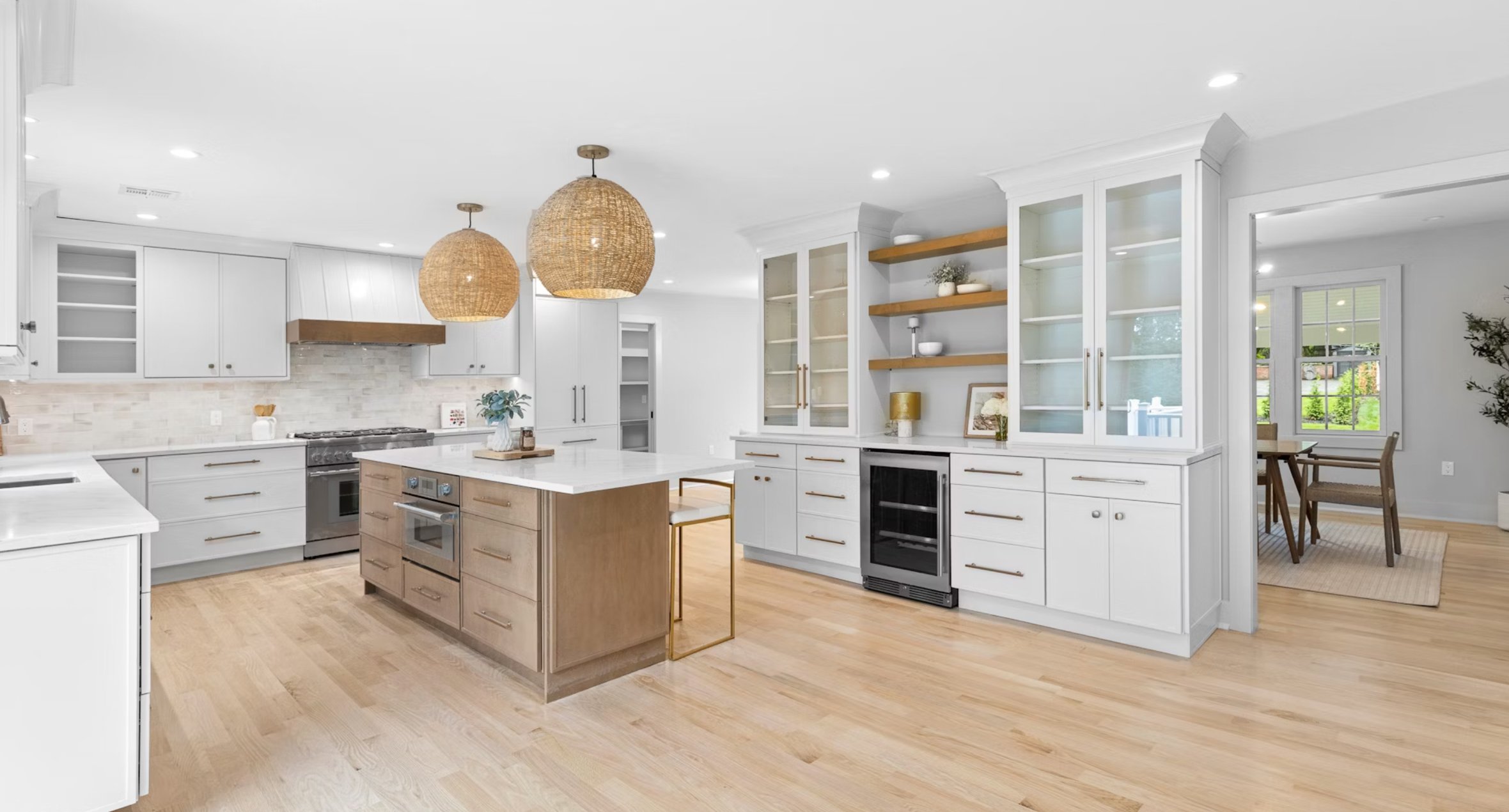 Bright, modern kitchen with white cabinets, a central island with wooden drawers, and two woven pendant lights, featuring hardwood flooring, open shelving, and a dining area visible through an open doorway.