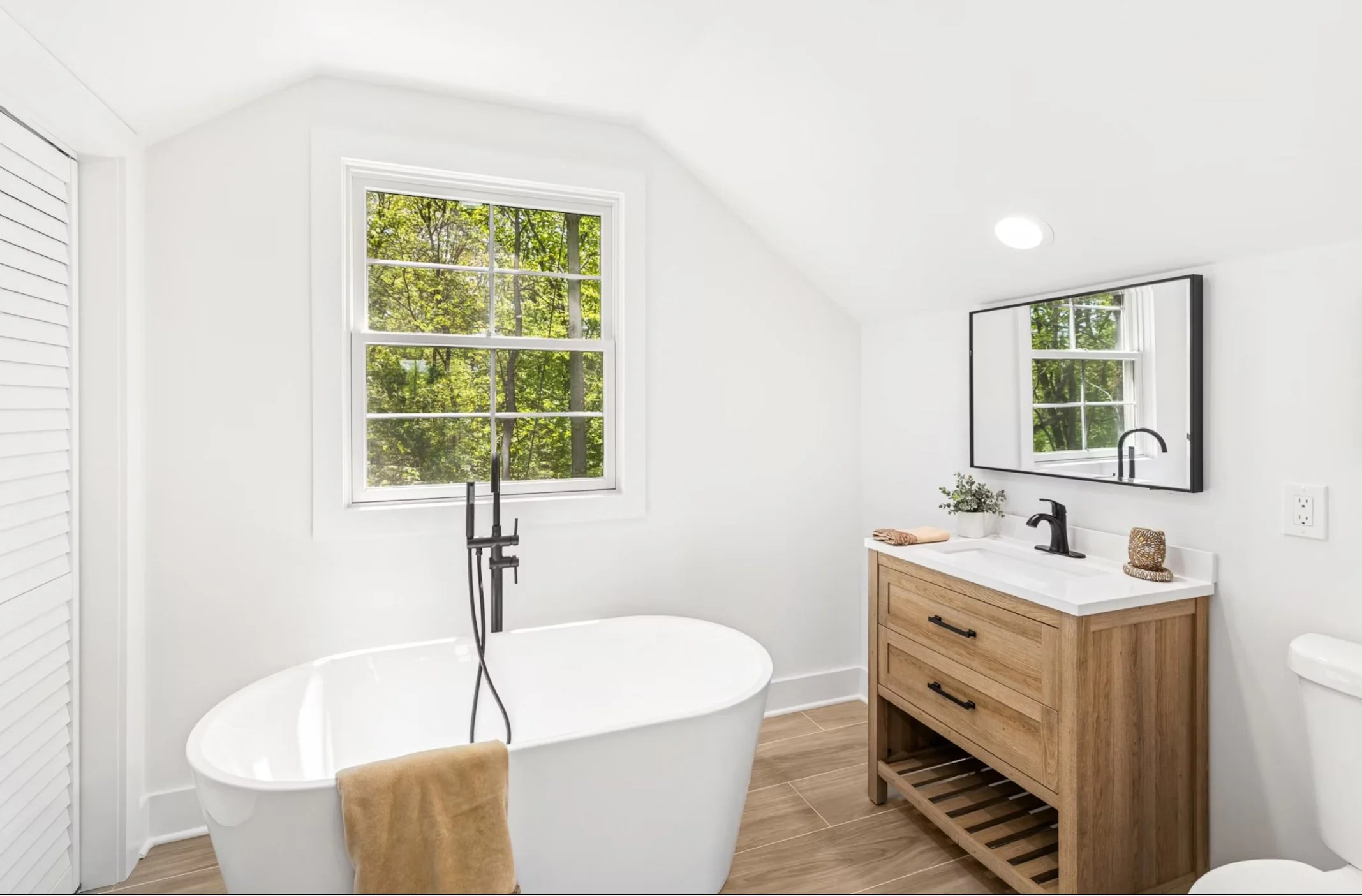 Modern bathroom with white walls, wooden vanity, large mirror, bathtub, window with green trees outside, and wooden floor.